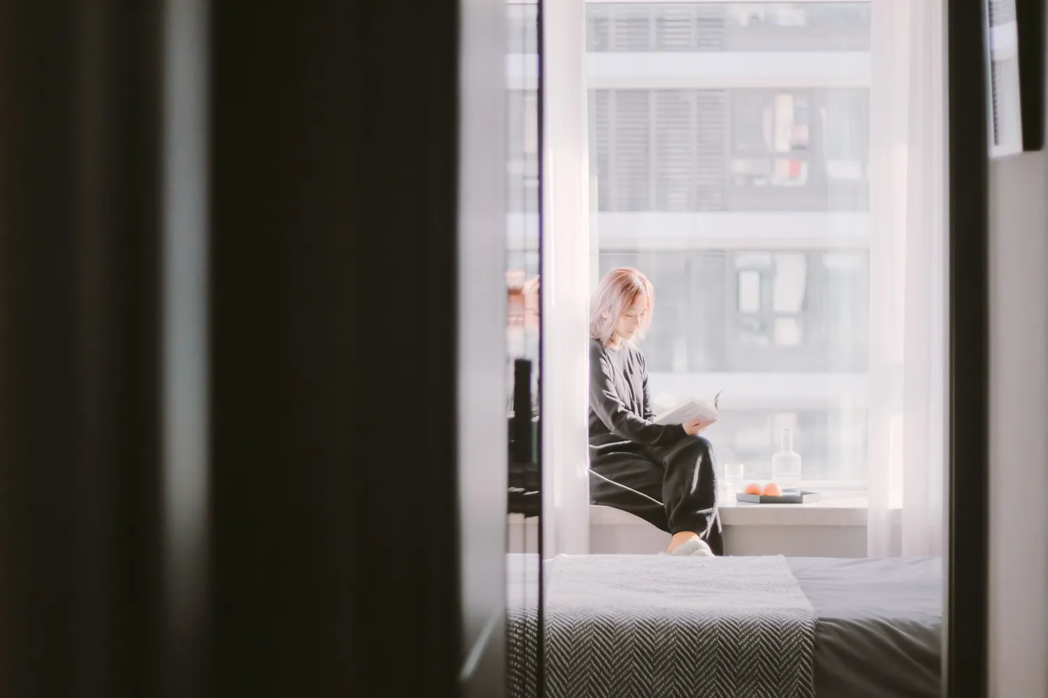 A woman with pastel-colored hair sits cross-legged on a window sill in a minimalist interior, reading a book with a glass of