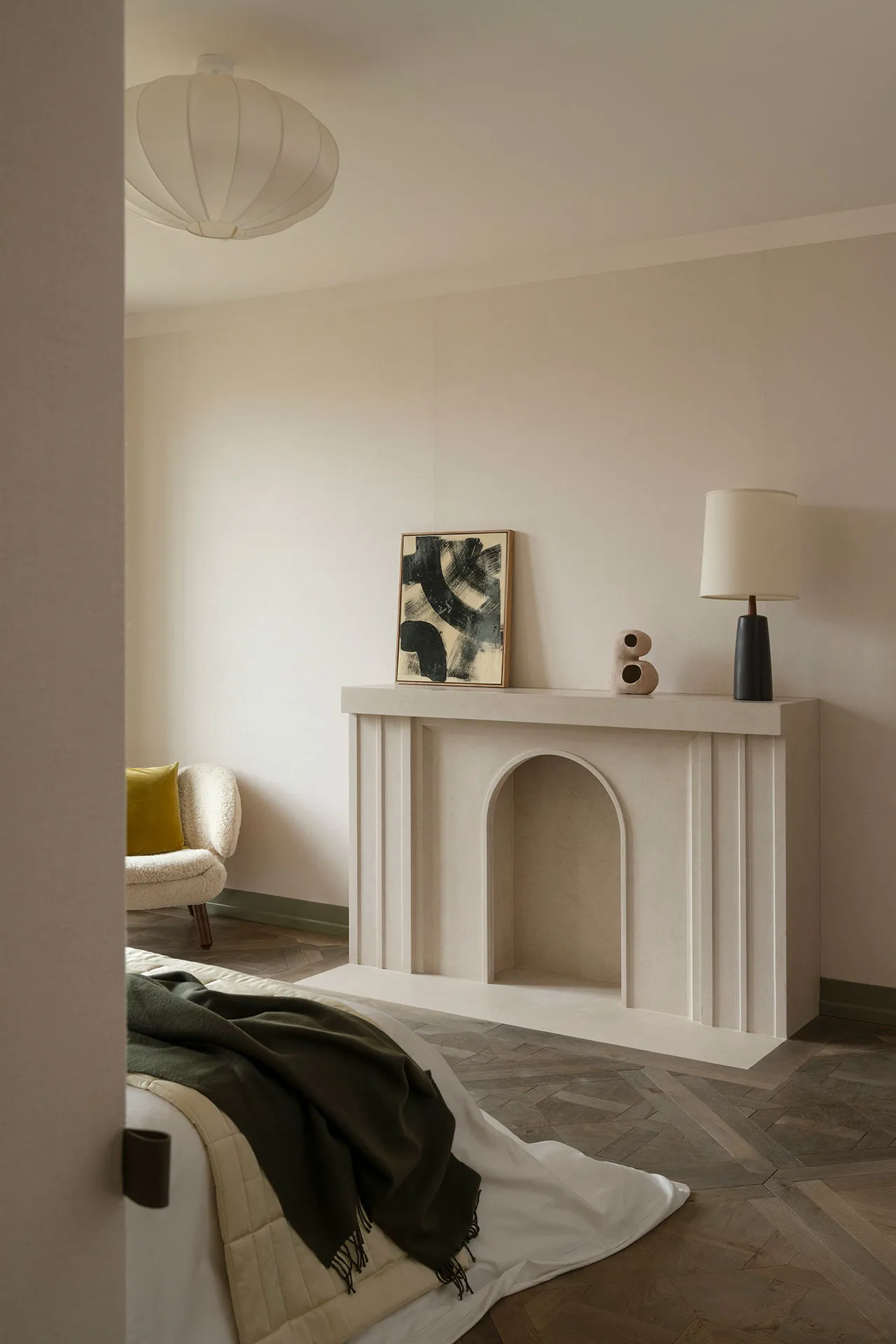 A minimalist bedroom featuring a concrete fireplace arch, a mustard yellow pillow, a black and white abstract painting, a lam