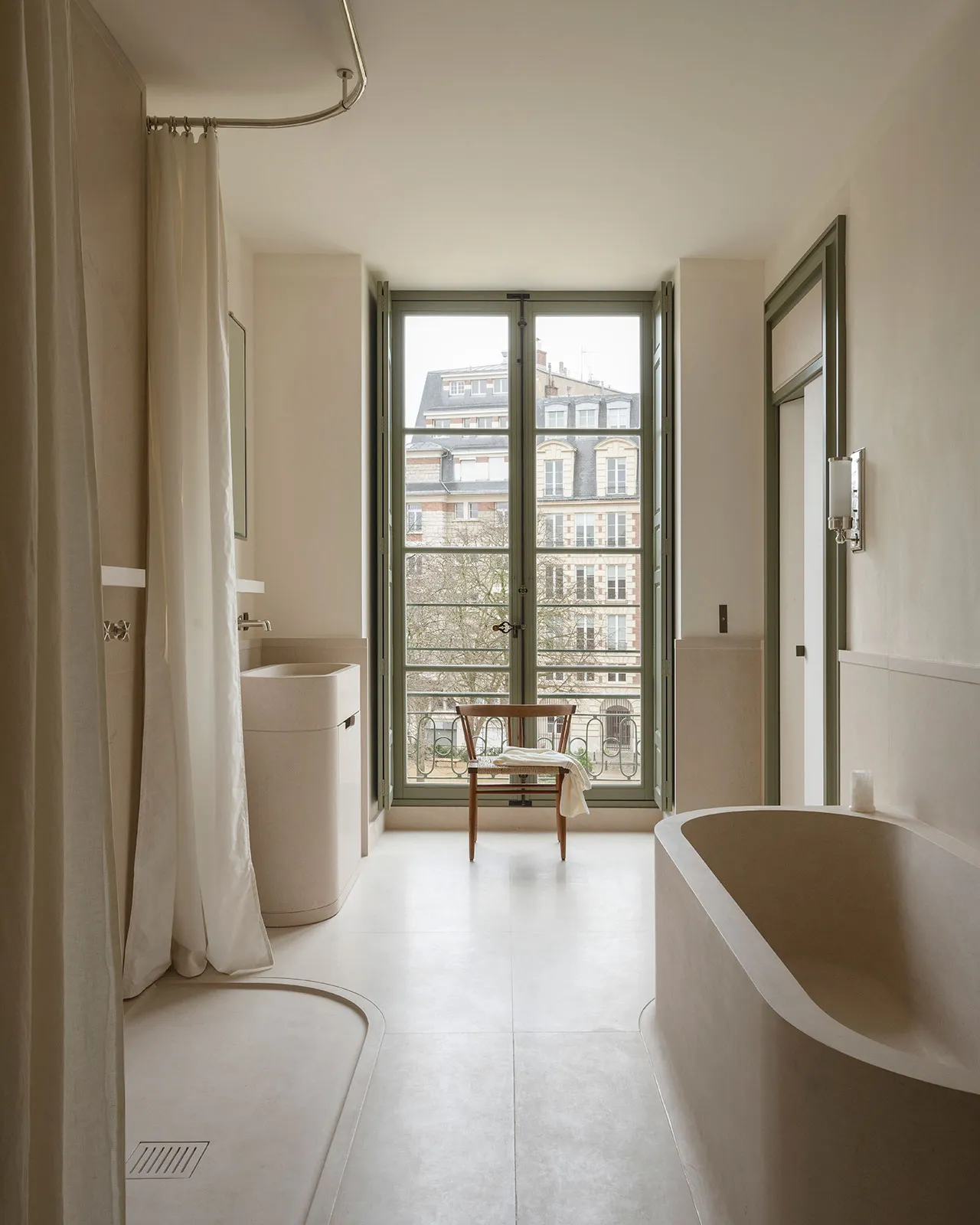 A modern minimalist bathroom with a concrete bathtub and flooring, large windows with a view of the city, and a simple chair.