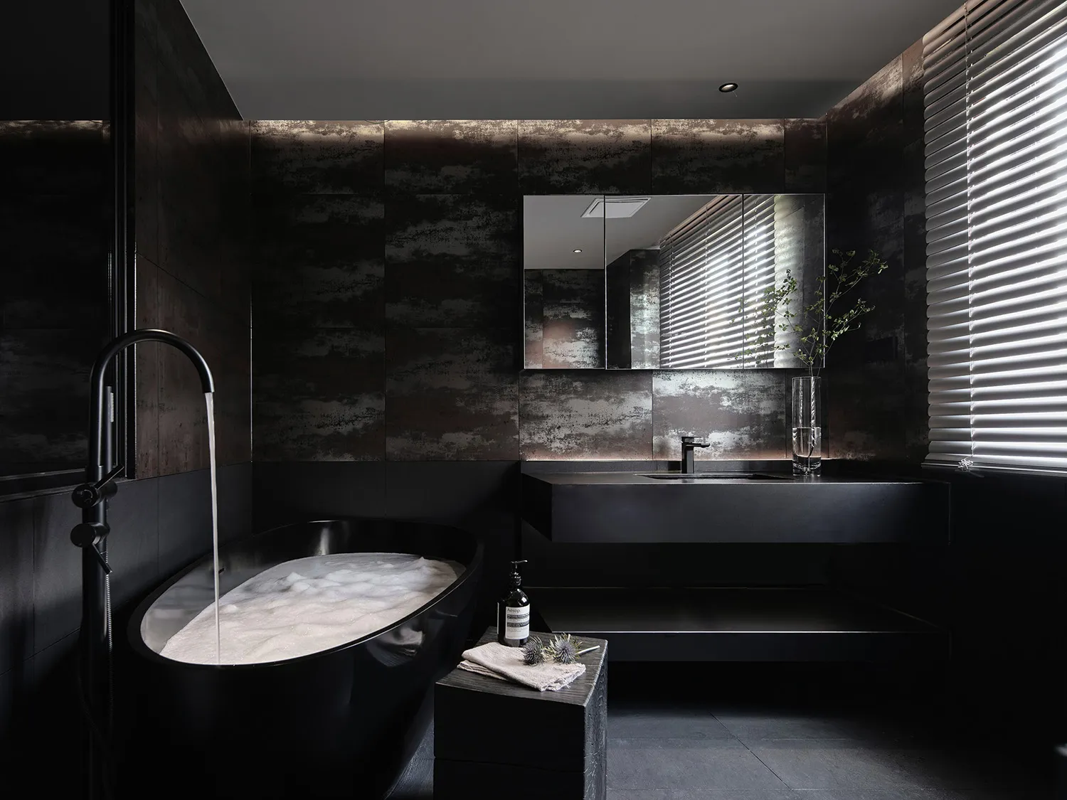 A minimalist modern bathroom featuring black tiles and concrete surfaces with a freestanding bathtub filled with bubbles unde