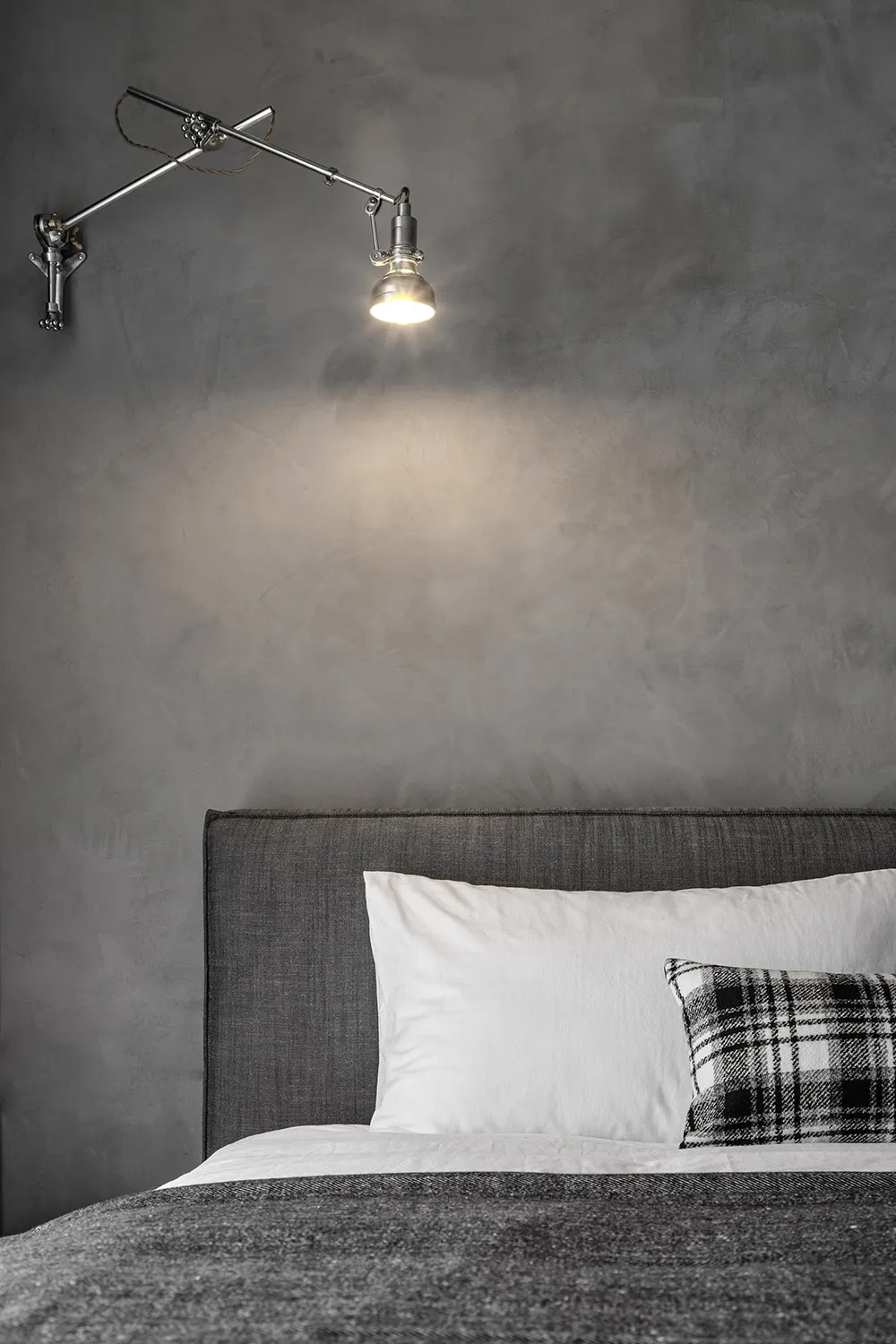 Industrially styled bedroom with concrete walls, a metal adjustable arm lamp, and a grey upholstered bed.