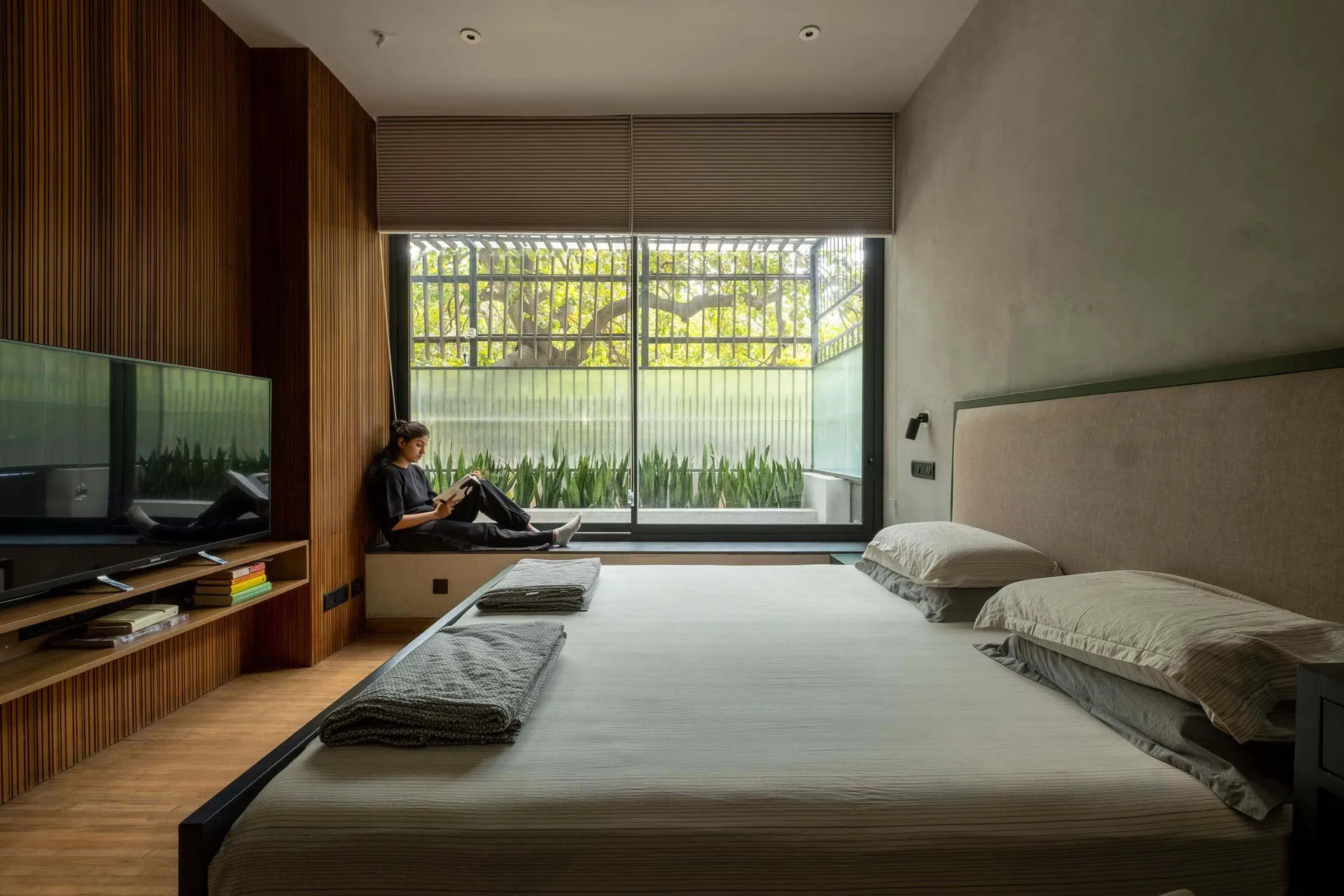 A modern minimalist bedroom featuring a large bed, wooden walls, and fabric bedding, with a person sitting on a window sill r