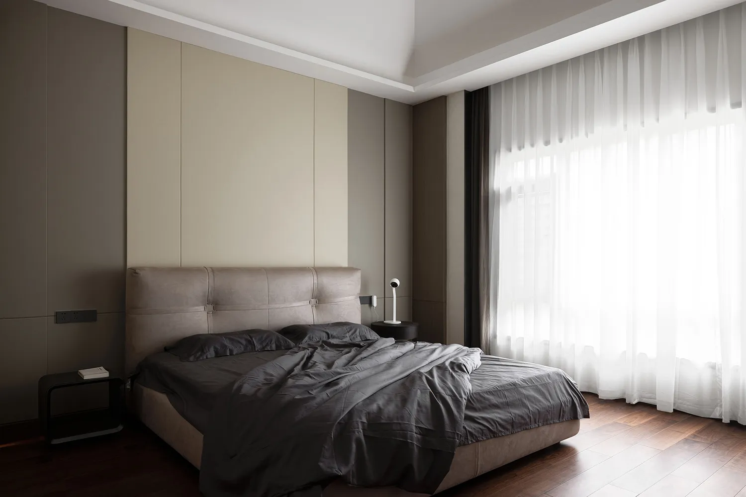 A modern bedroom featuring a brown upholstered bed with grey bedding and curtains, set against a muted color palette with a l