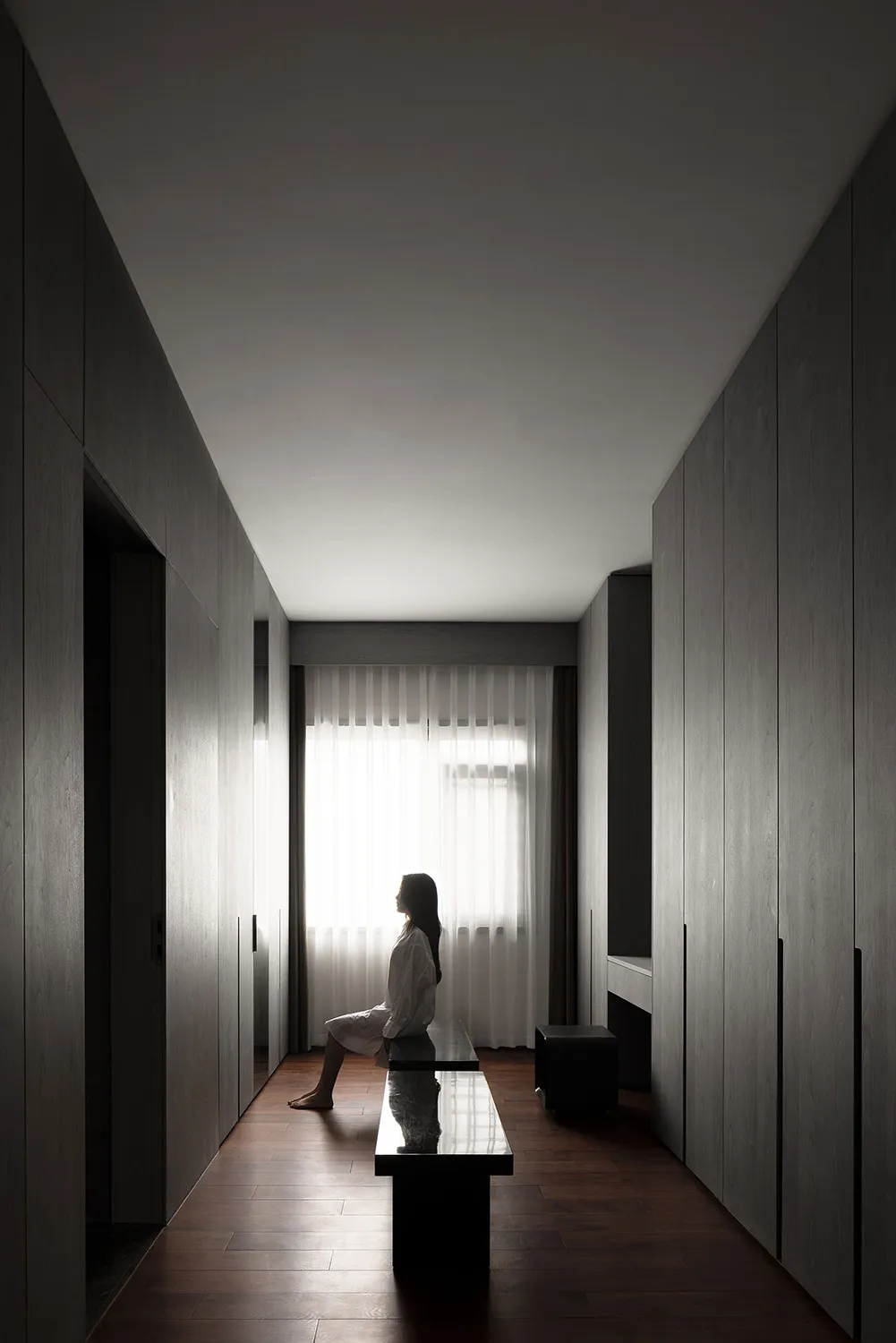 A minimalist interior space with wood elements, featuring a woman sitting on a bench by a window.