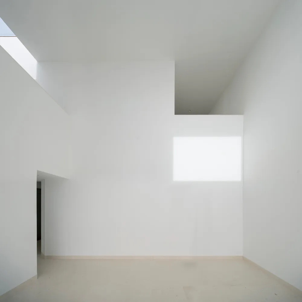 A minimalist white interior space with a high ceiling, light shining through a window, and a clean, uncluttered design.