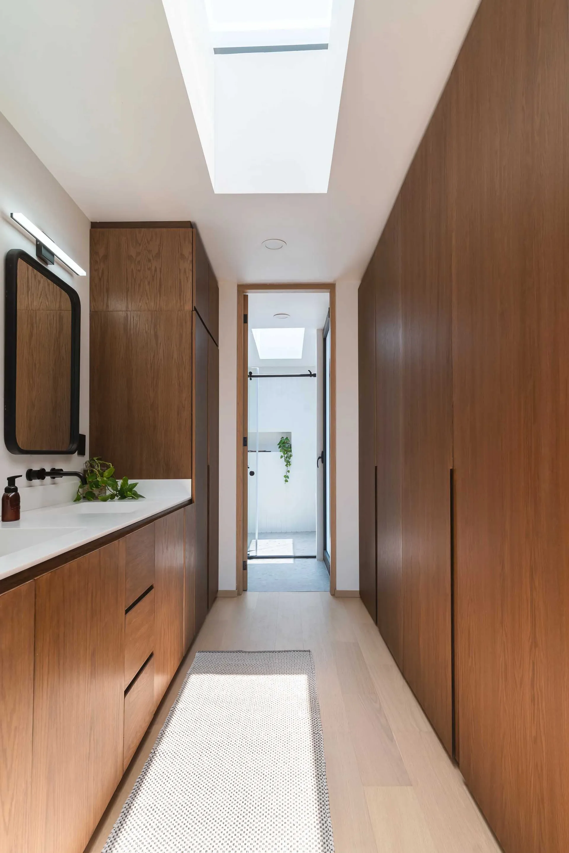 Modern bathroom with wooden cabinets, white flooring, and a skylight above a long corridor leading to a shower stall.