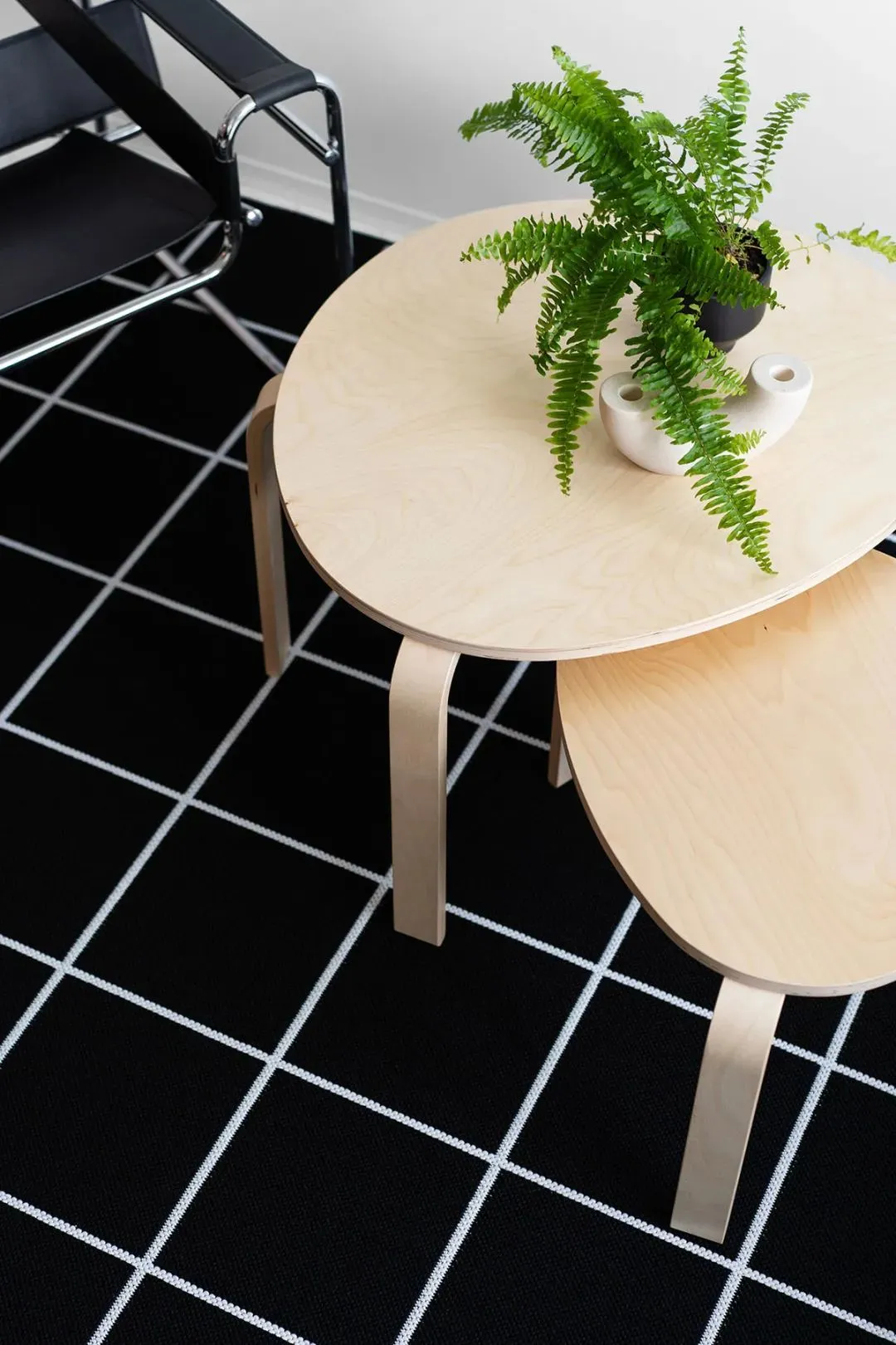 Two minimalist, round, wooden side tables stacked, with a green fern plant and a white ceramic dish on the larger table.