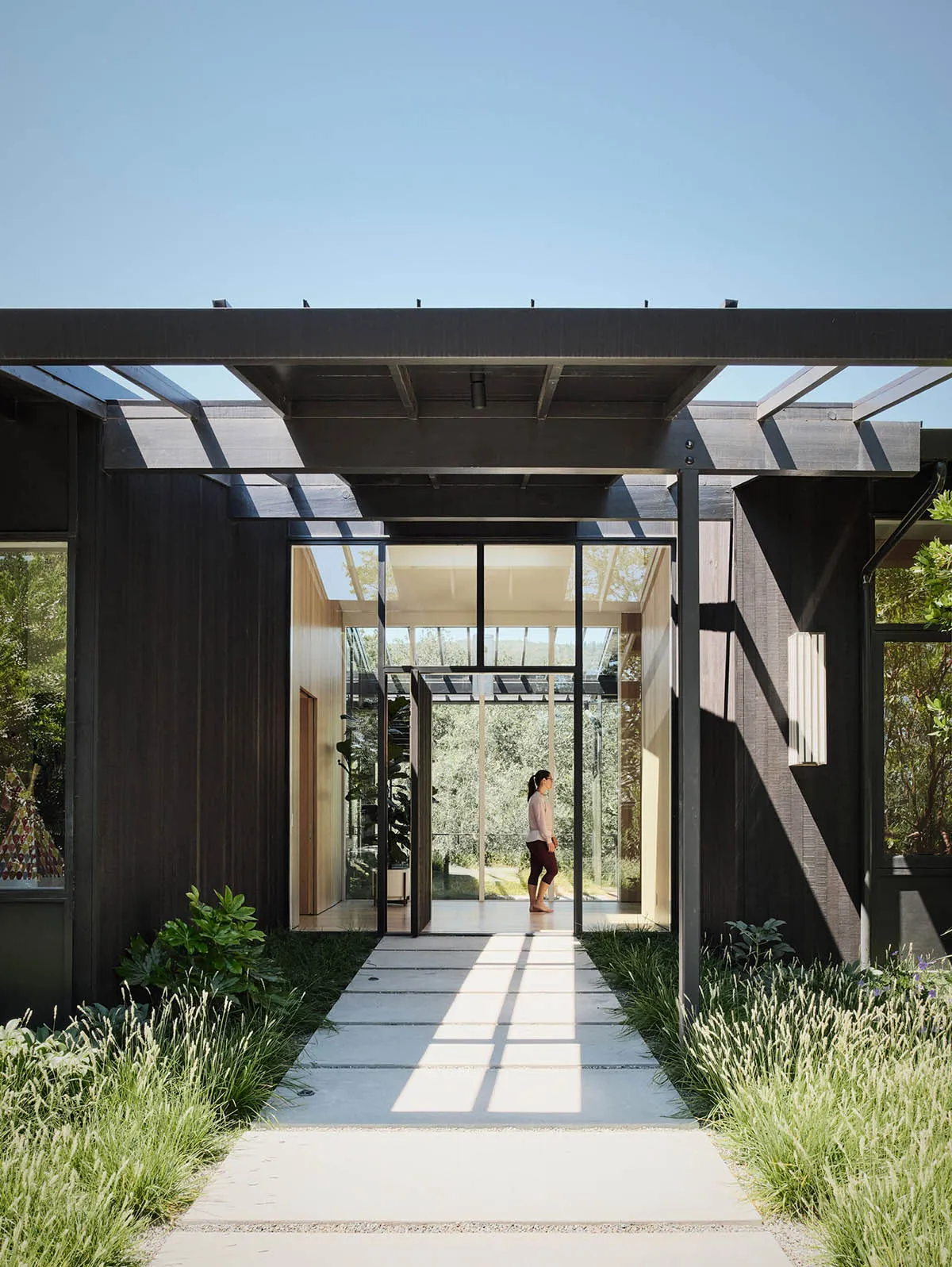 Modern house exterior with large glass windows and wooden accents, featuring a pathway and greenery.