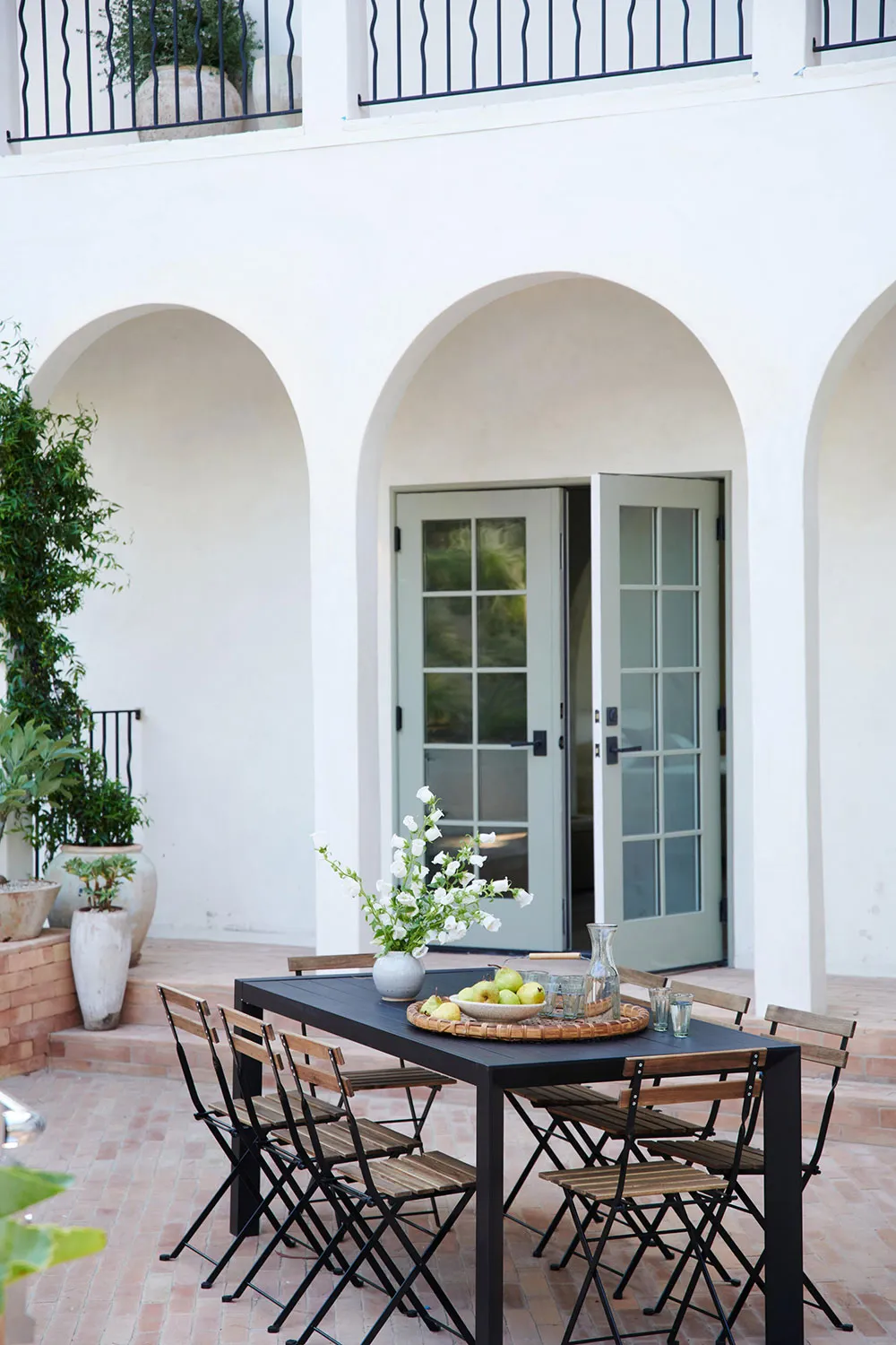 Outdoor dining area with a black metal table, wooden chairs, white flowers, and a fruit bowl.