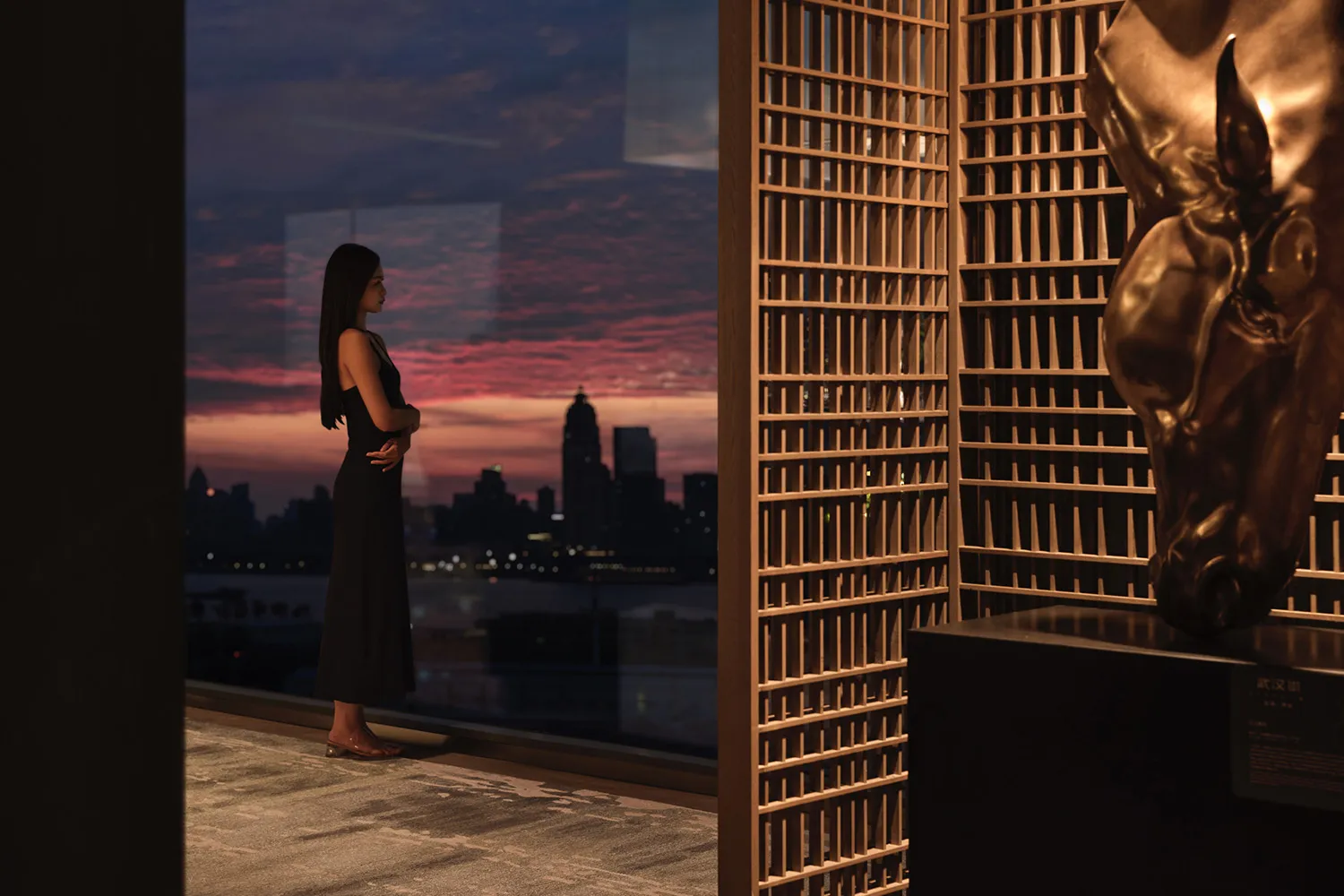 A woman standing on a wooden floor with a steel railing, observing a cityscape at sunset through a large window.