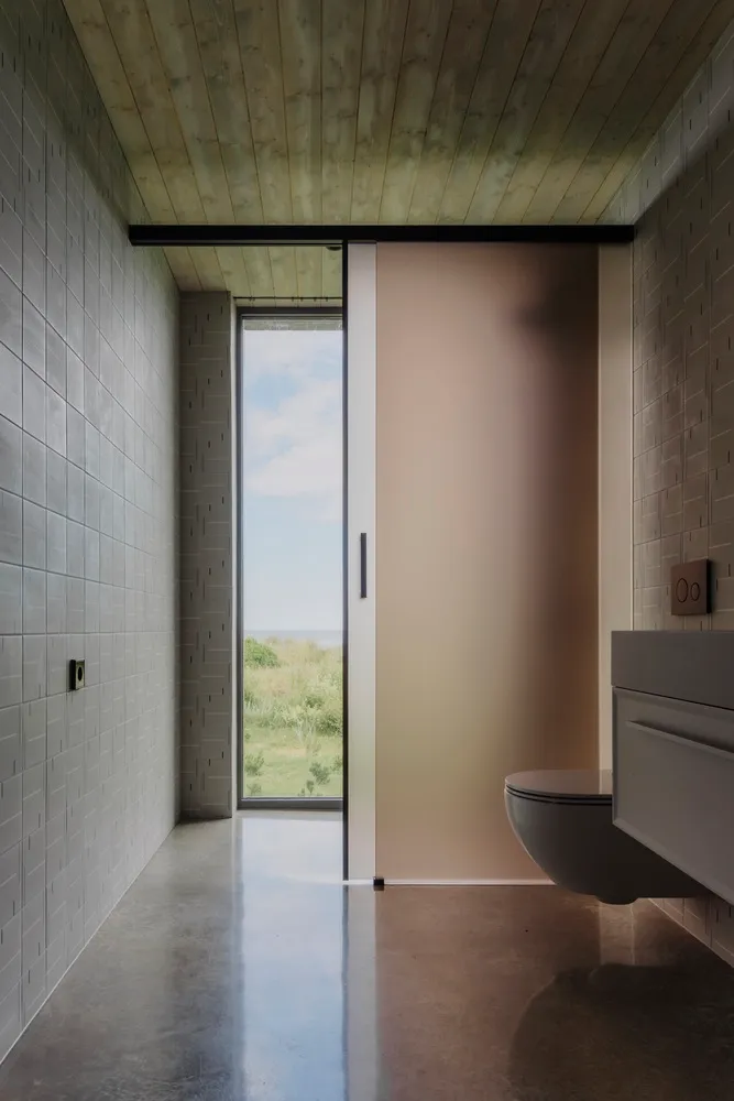 Modern industrial-style bathroom with concrete floor and wood paneling, featuring a frosted glass sliding door and a window o