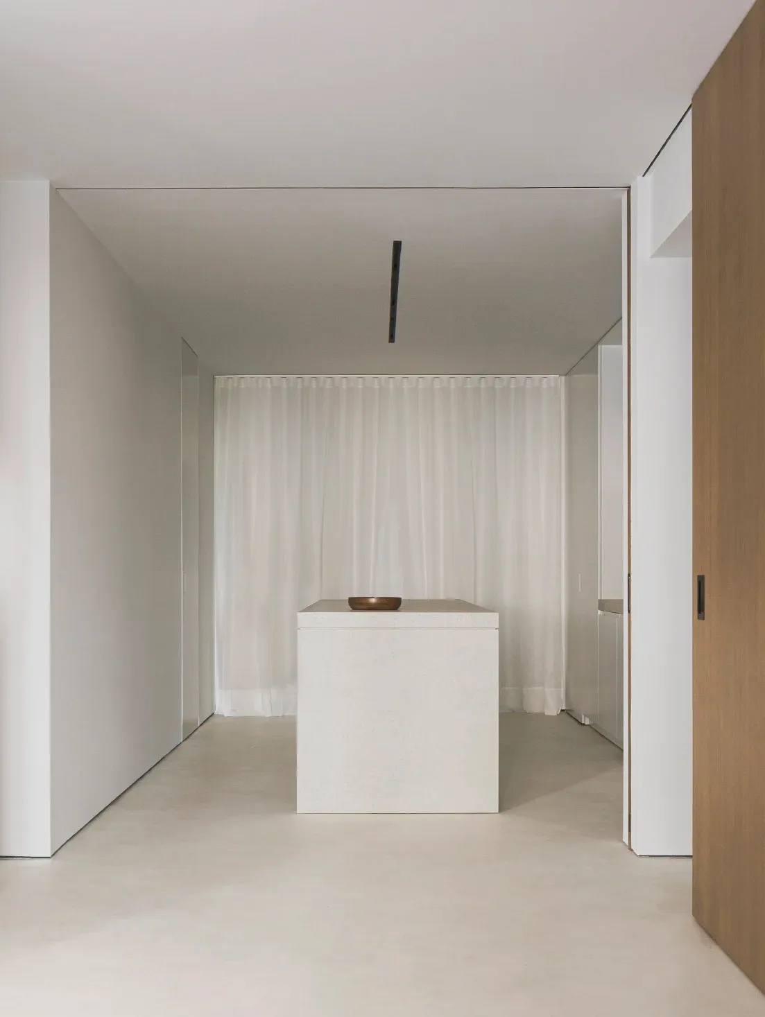A minimalist interior design featuring a concrete cube with a wooden bowl on top in the foreground, with sheer curtains and w