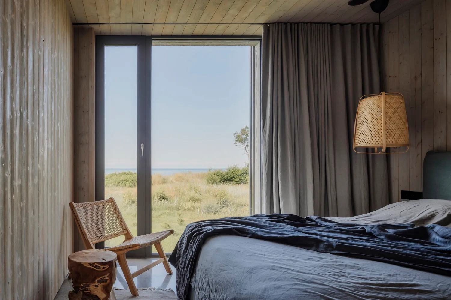 A serene minimalist bedroom with a large window overlooking a natural landscape, featuring a wooden bed, chair, and stool, an