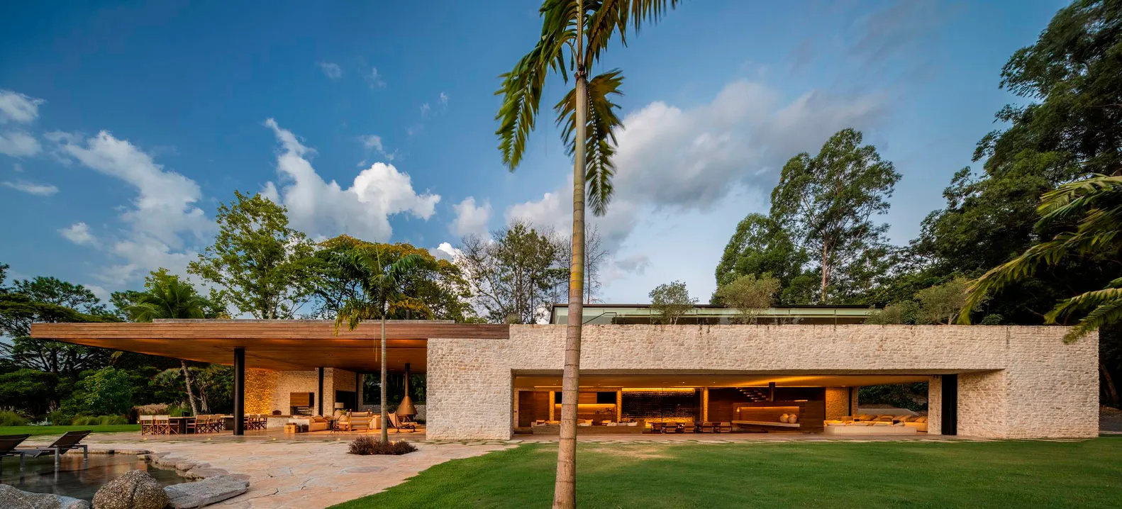 A modern house with a sandstone exterior, wooden roof, and glass walls surrounded by a tropical landscaped yard.