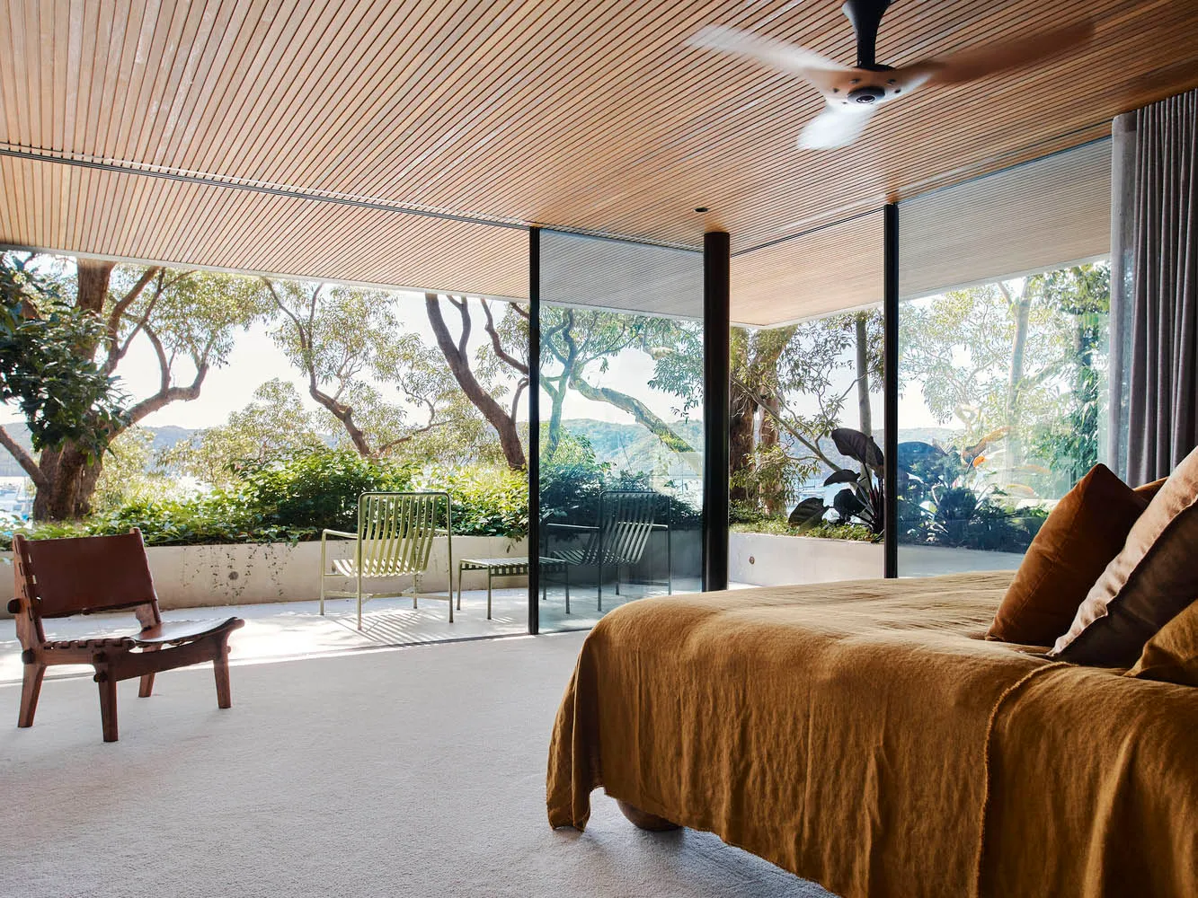 Modern bedroom with wooden ceiling and floor, featuring a large bed, a chair, and a view of trees and a garden.