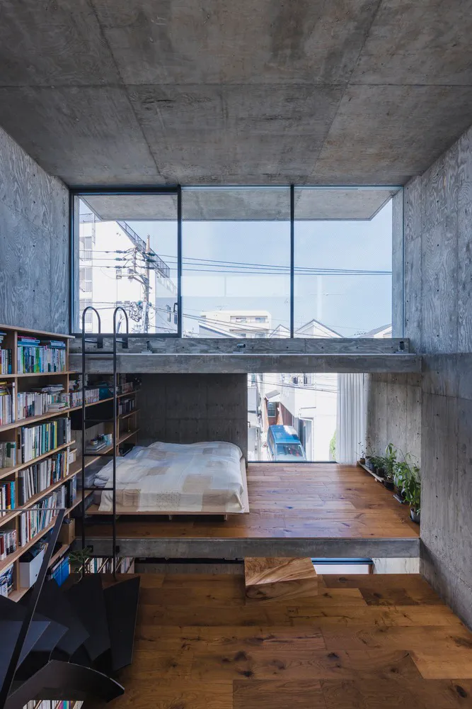A minimalist bedroom featuring a concrete floor and wall, with a bed on a raised platform and a bookshelf.