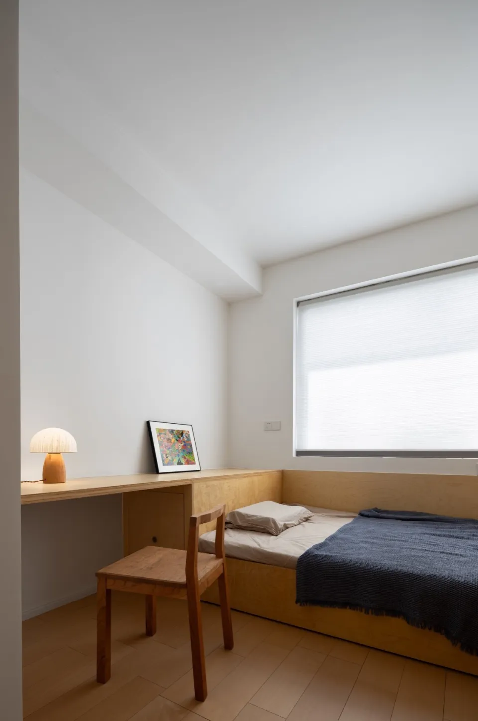 Modern minimalist bedroom with a solid wood bed, desk, and chair, featuring a large window with blinds.