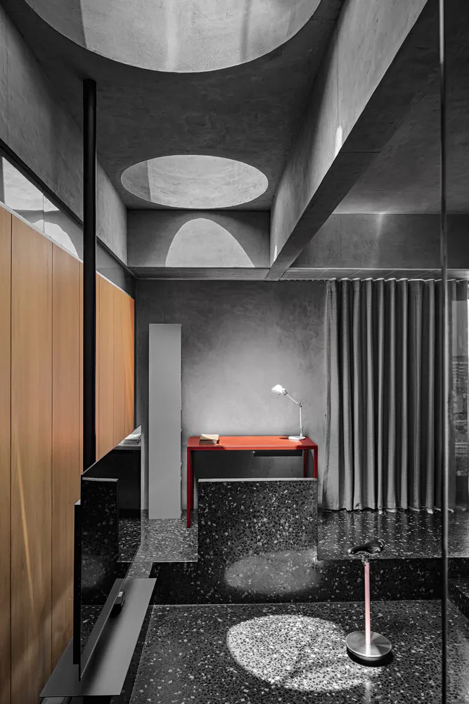 A minimalist office workspace featuring a red desk with a lamp, placed on a terrazzo floor, with curtains and a concrete ceil