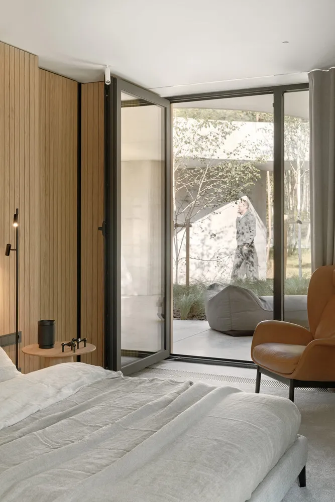 A modern bedroom with a white bed, wooden walls, and open doors leading to a garden.