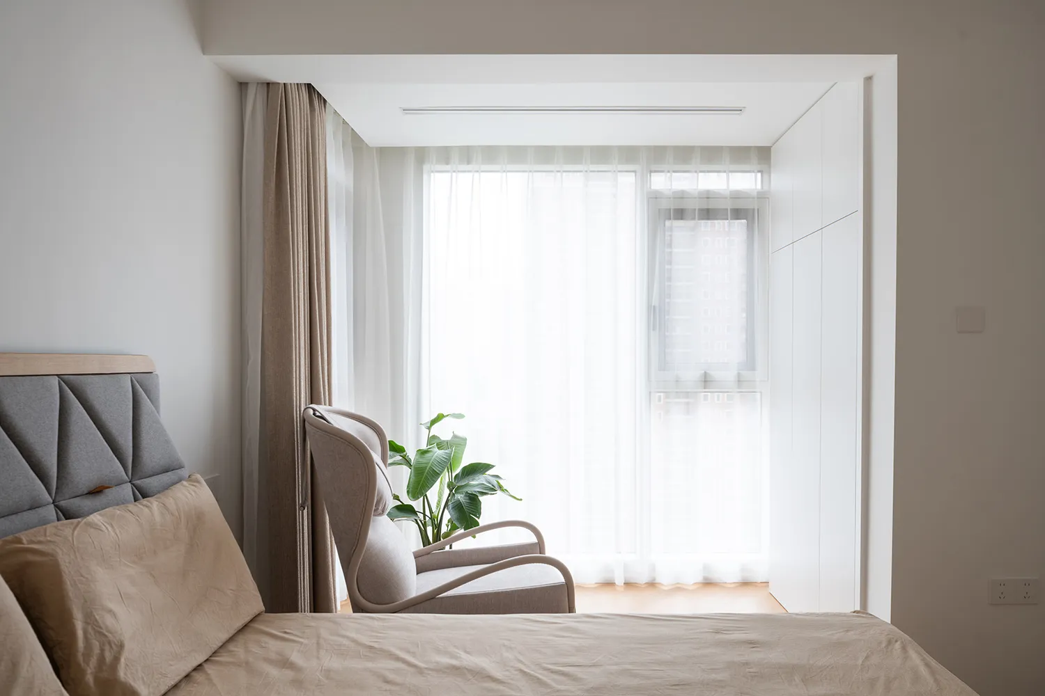 A minimalist bedroom featuring a beige upholstered bed, a modern beige armchair, light wood flooring, and sheer curtains on a