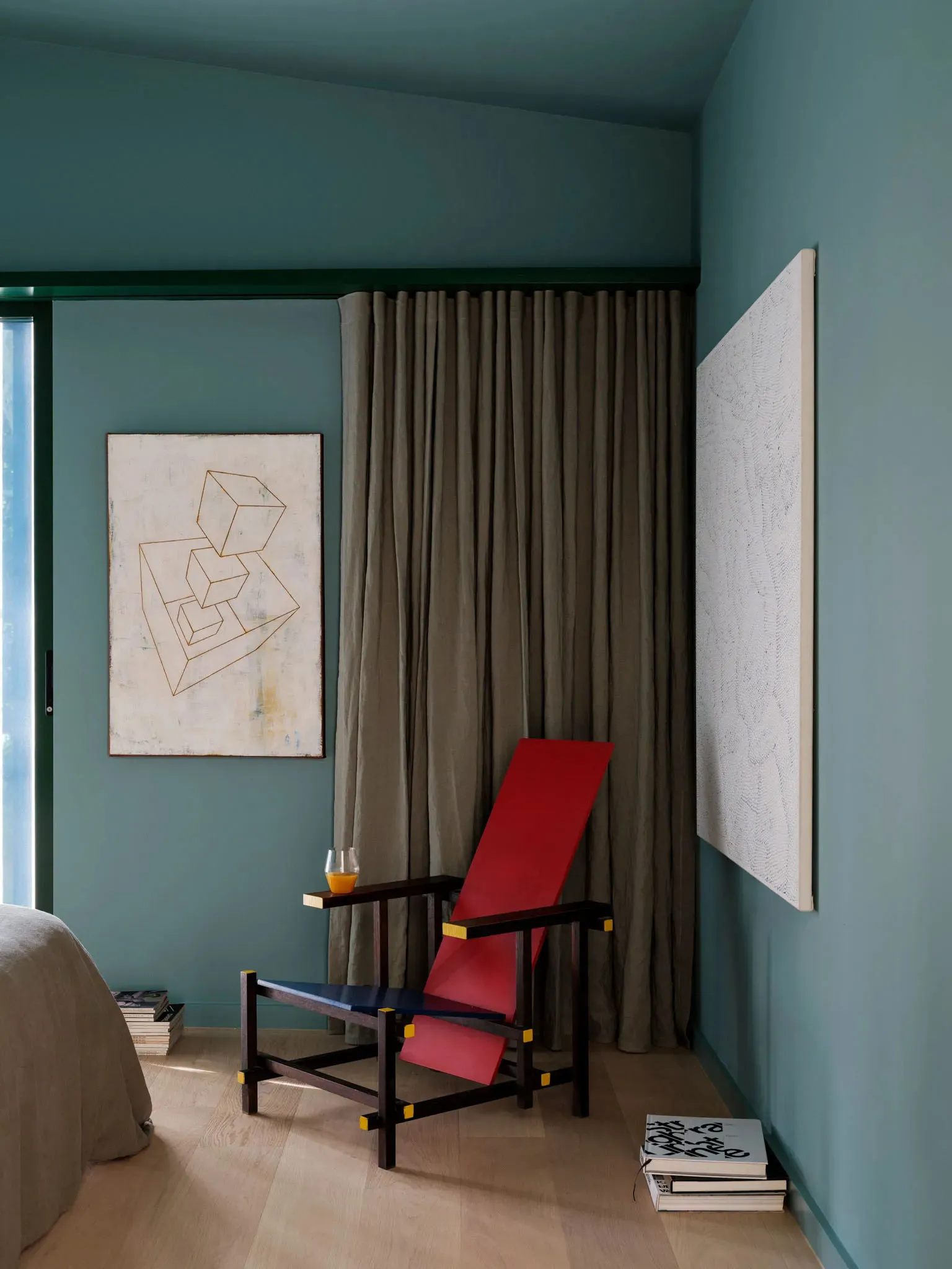 A modern bedroom featuring fabric curtains, a vibrant red and blue reclining chair, and geometric abstract art on the walls.