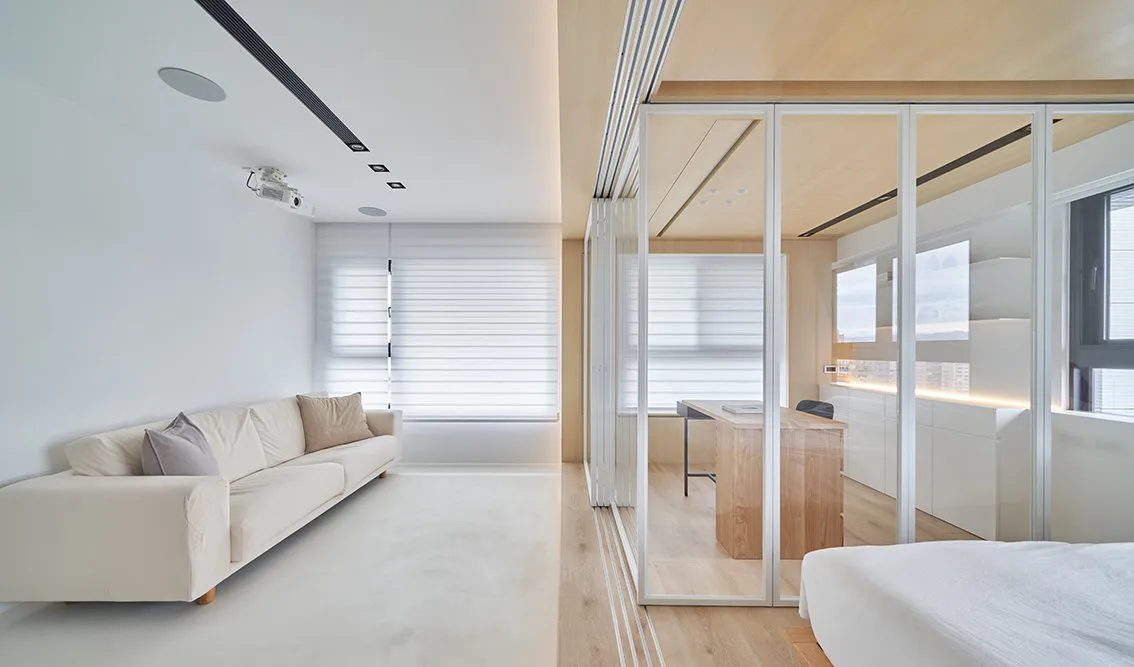 A modern living room with a neutral color palette featuring a comfortable sofa, blinds, and a study area with a desk and wind