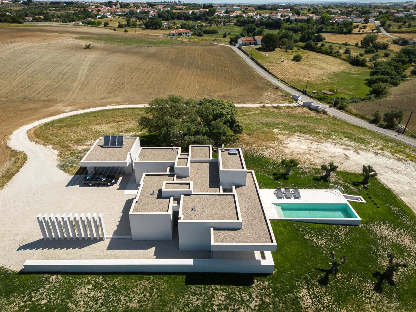 Aerial view of a modern villa with concrete finishes, featuring a swimming pool and a carport in a rural area.