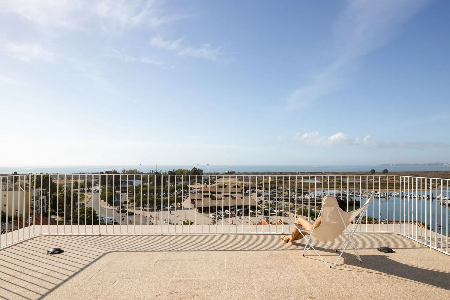 A person sitting in a white chair on a balcony with a metal railing overlooking a coastal landscape.
