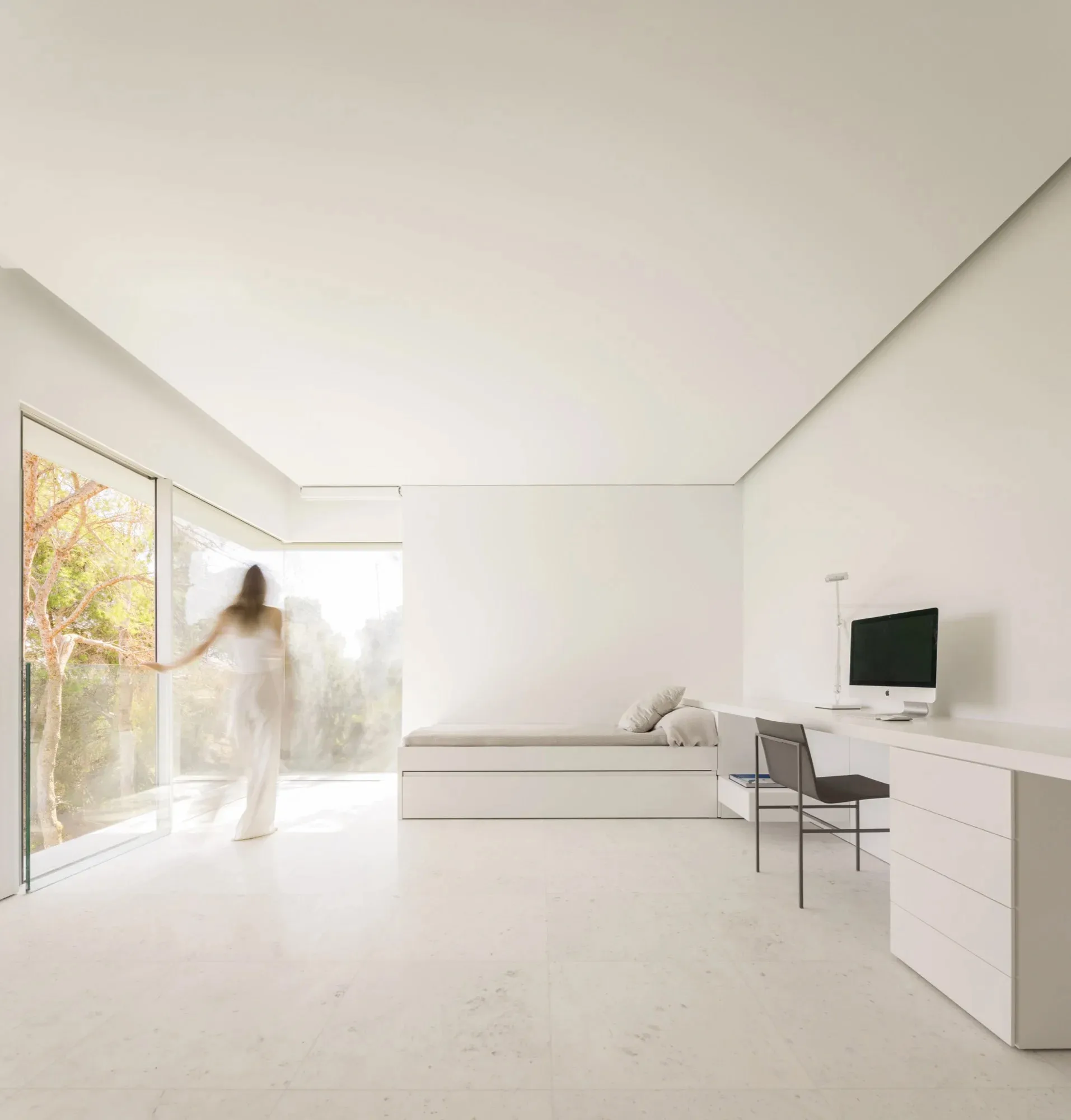 A minimalist bedroom featuring a sleek, modern bed with integrated storage, a sleek white desk, and large windows with glass
