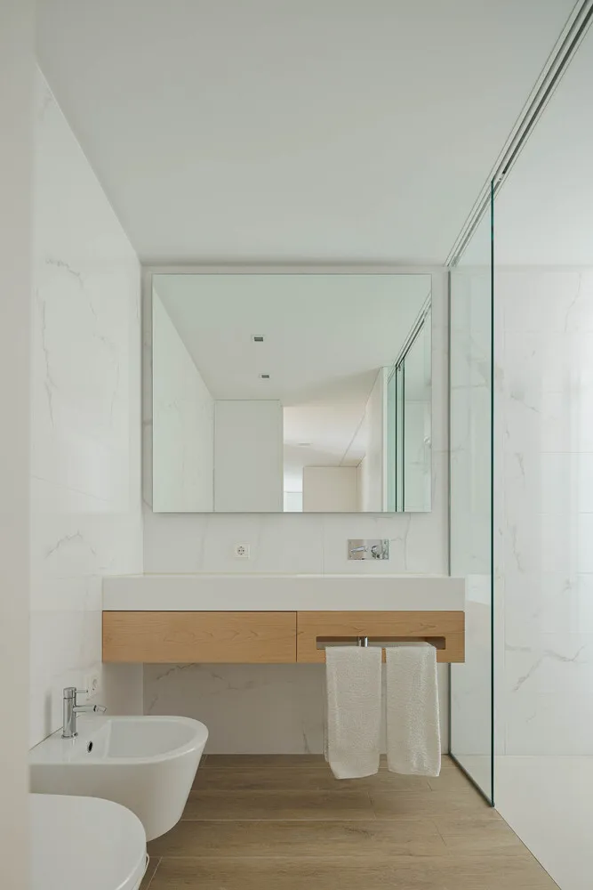 Modern bathroom featuring white marble walls, a floating wooden vanity with a white countertop, and a glass-enclosed shower a