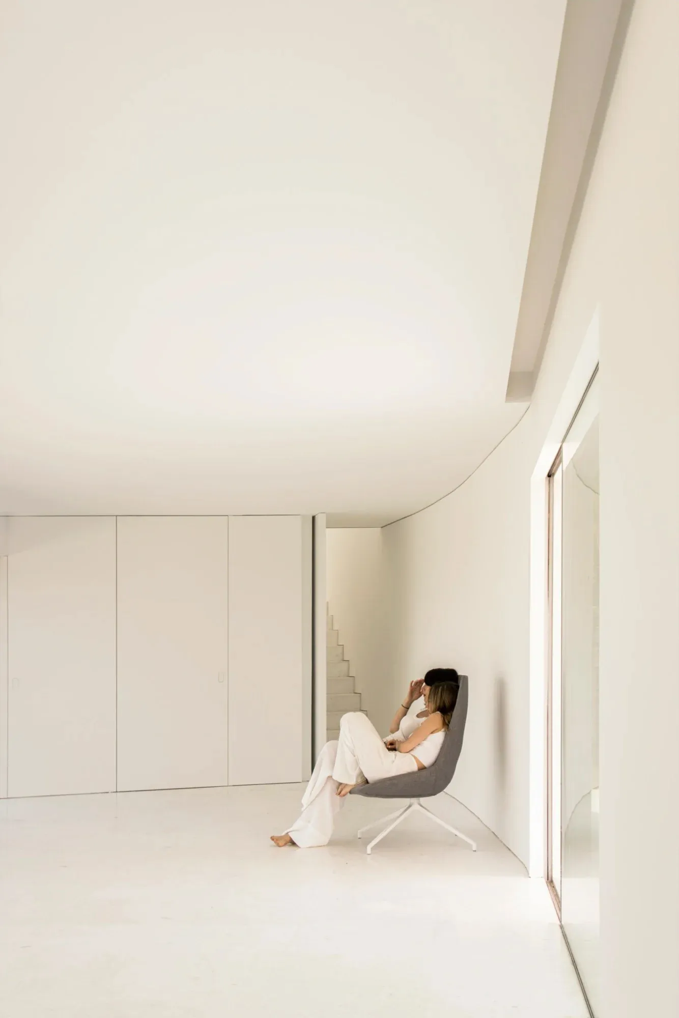 A minimalist living room with a white floor, white walls, and a person sitting on a gray chair.