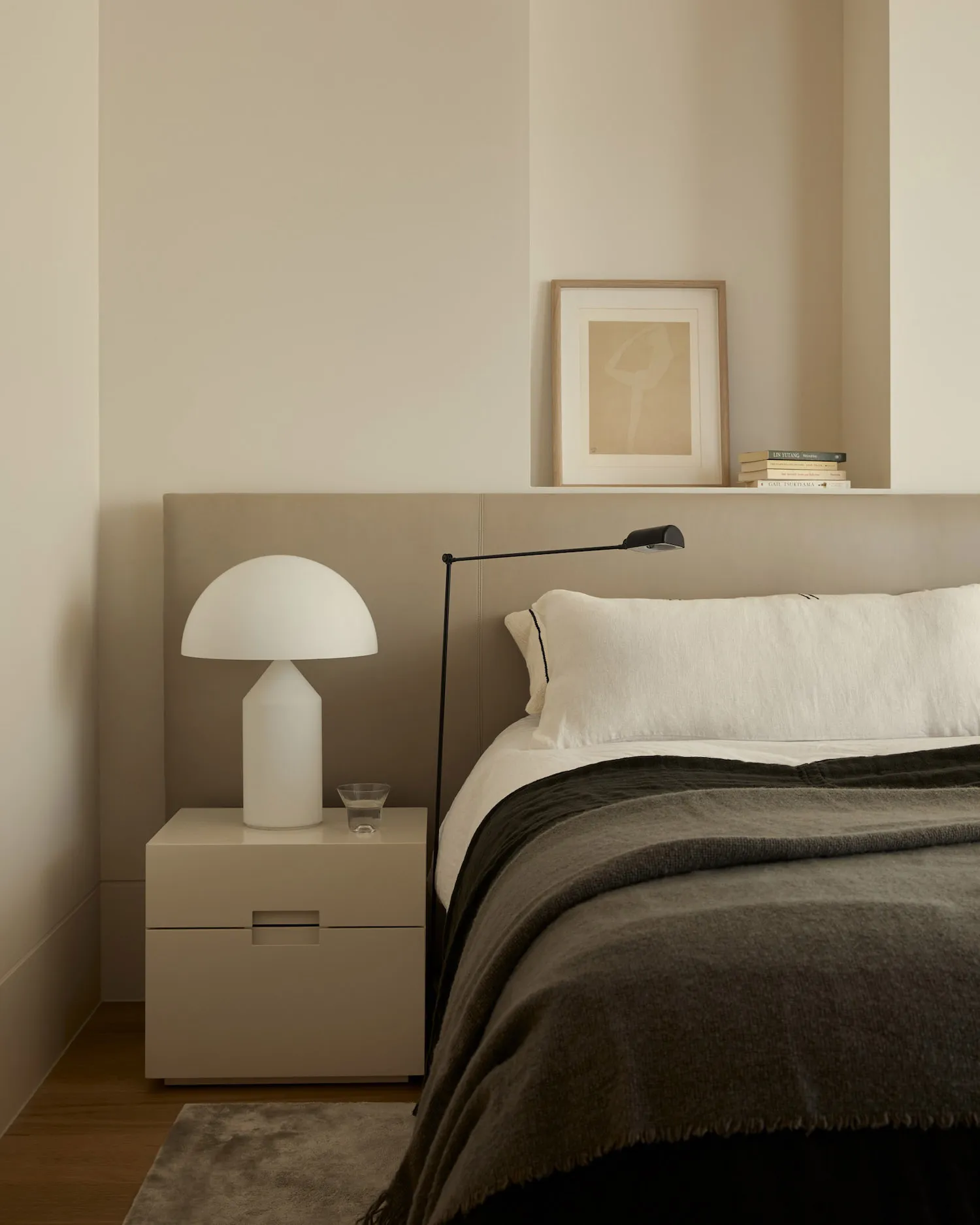 A minimalist bedroom with a white bed, a nightstand with a white lamp, and a floating shelf above the bed.