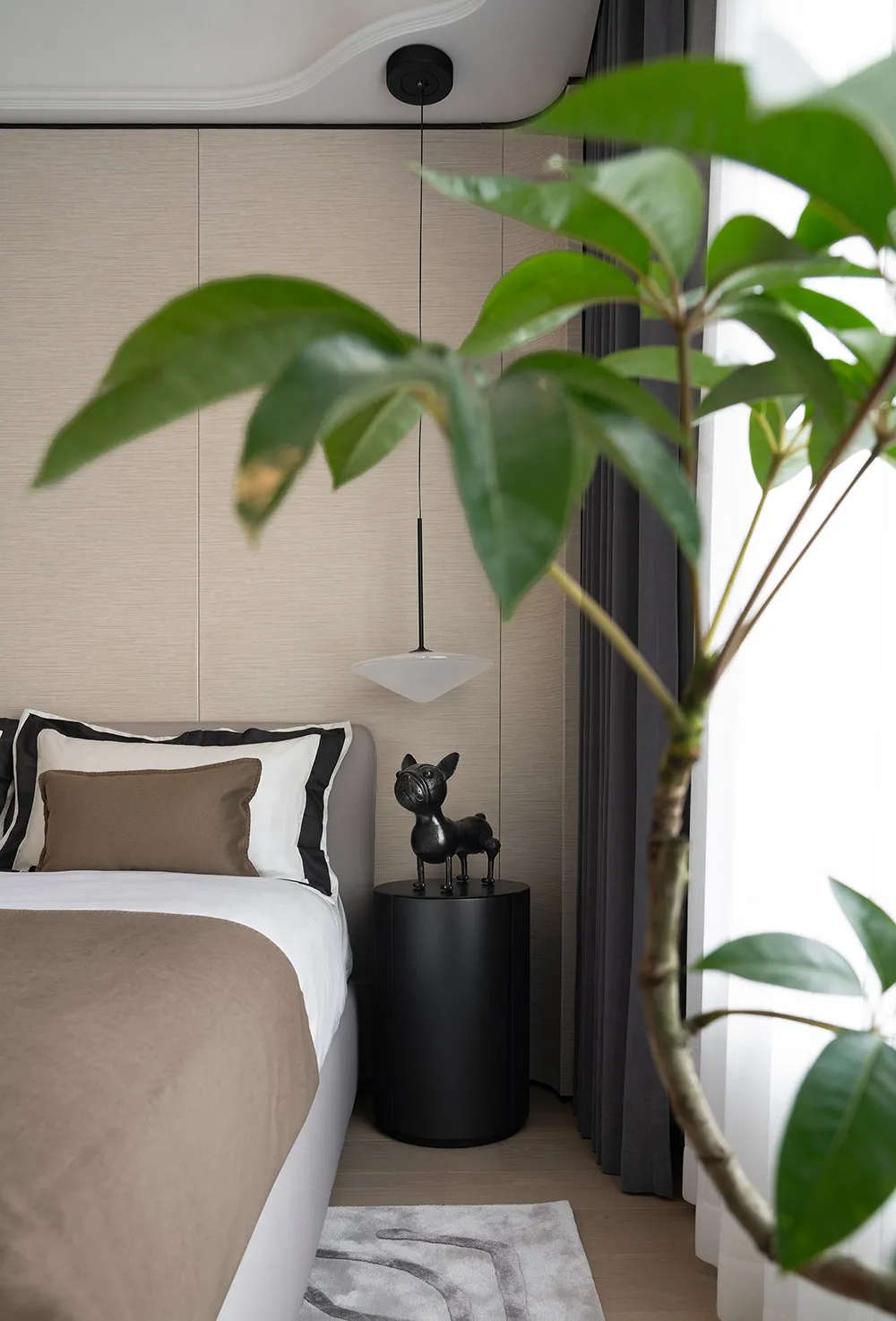 Modern bedroom featuring textured beige wall panels, dark curtains with white sheers, black bedside table, brown blanket, and