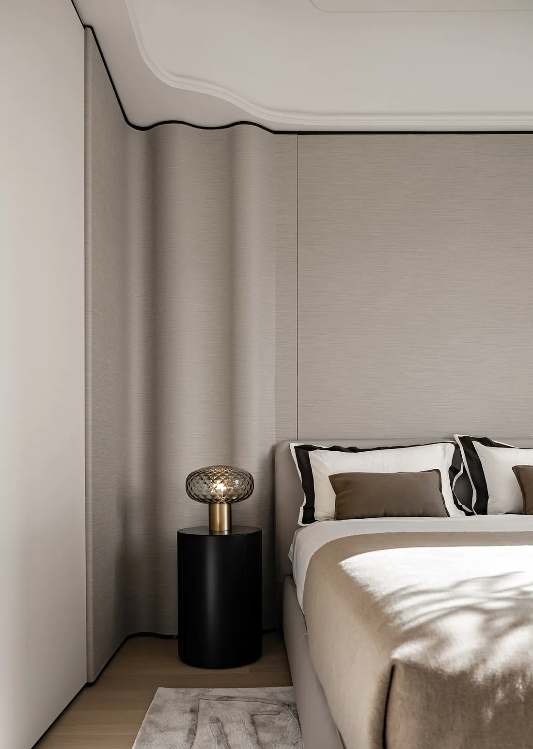 Modern minimalist bedroom featuring a bed with taupe fabric bedding and a side table with a decorative lamp.