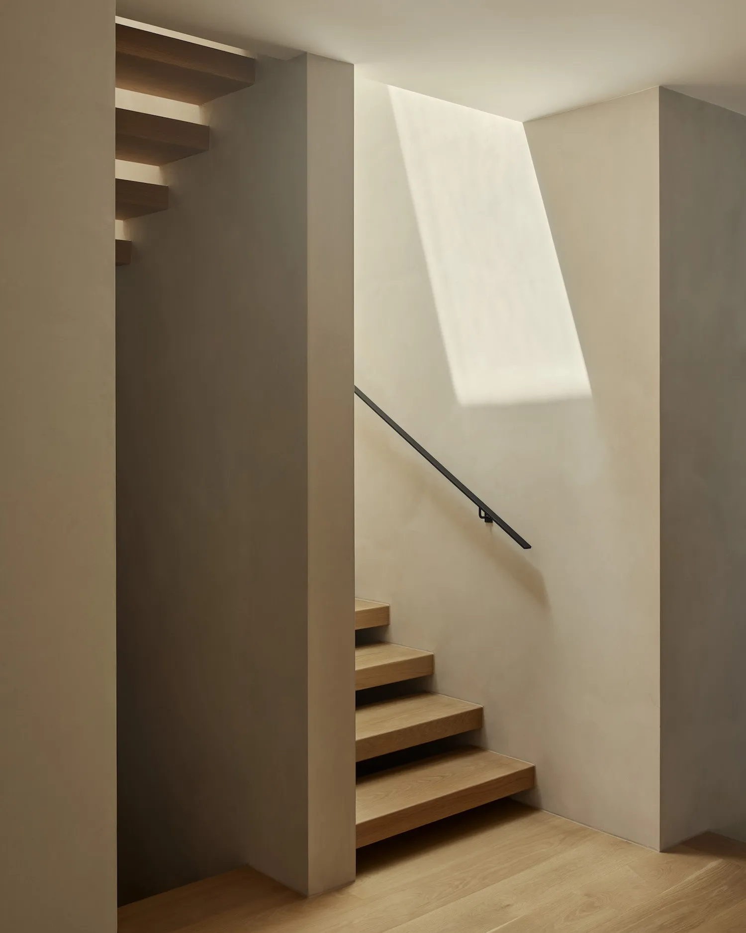 A modern minimalist staircase with a wooden platform and handrail. The staircase features a skylight allowing natural light t