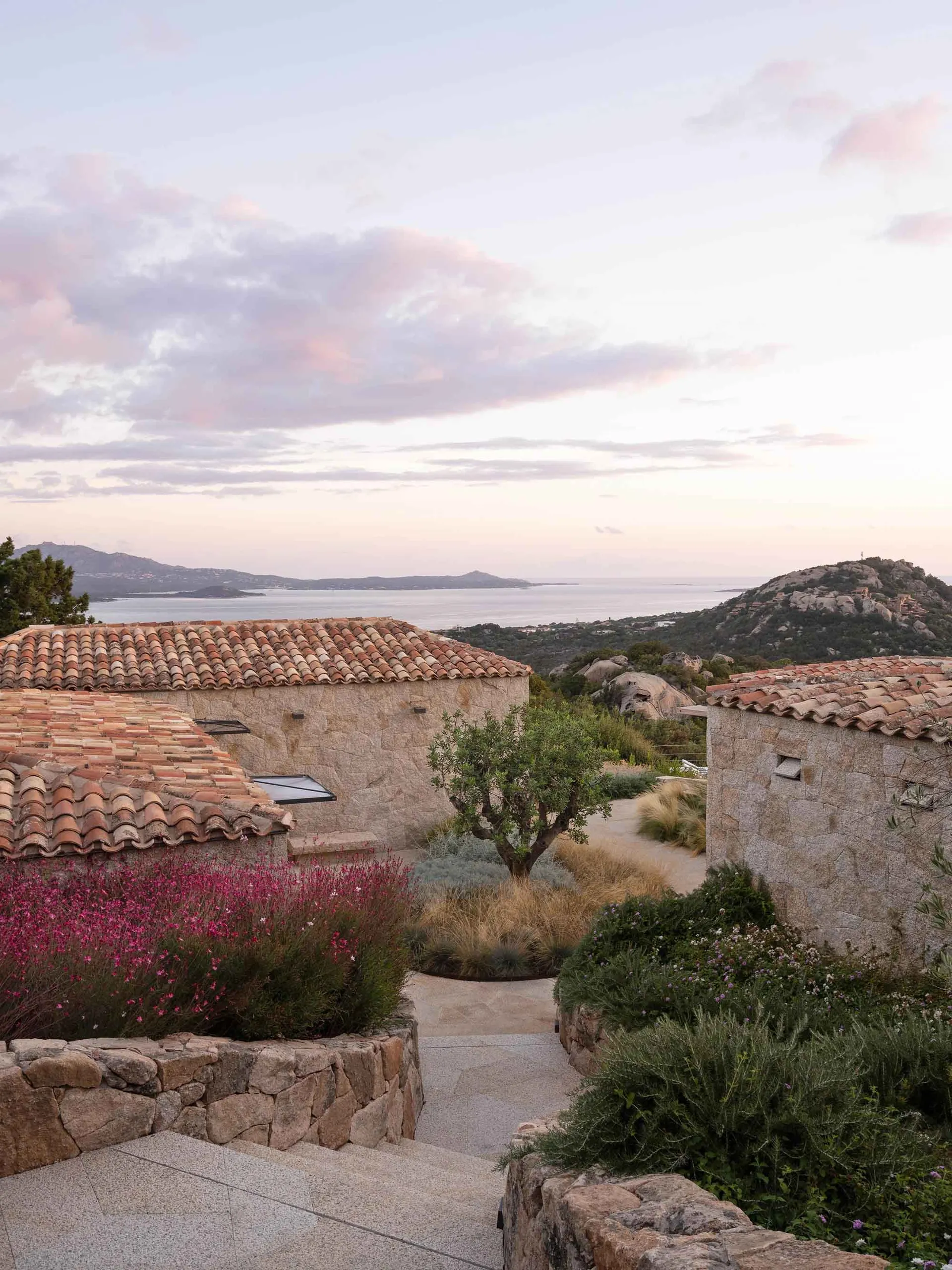 Rustic exterior of a residential house with stone walls and a terracotta roof, surrounded by lush vegetation and overlooking