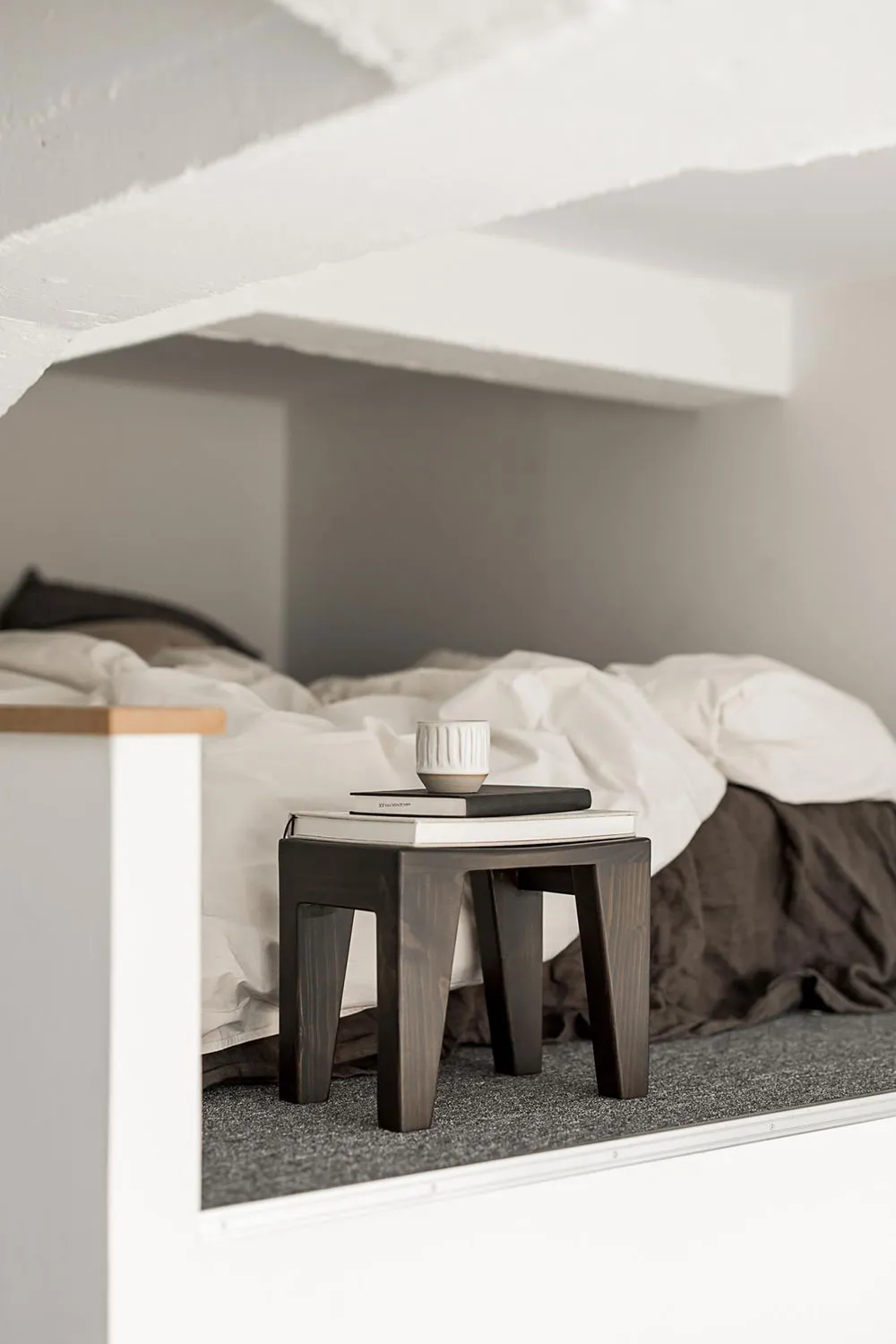 A minimalist bedroom featuring a wooden bedside table with books and a mug on top.