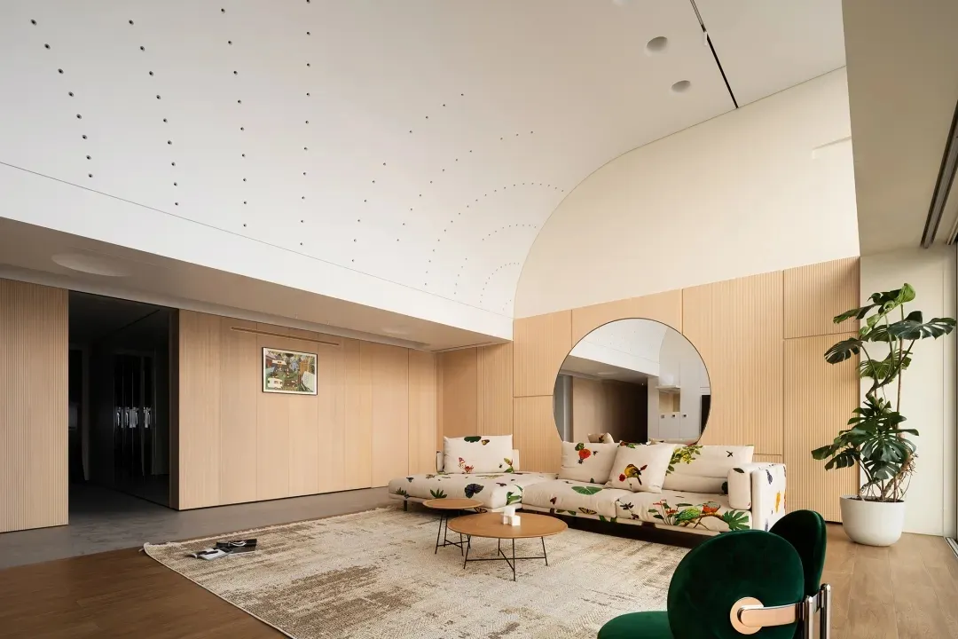 Modern living room featuring a wooden couch with botanical print upholstery, a large round mirror, and a green velvet chair.