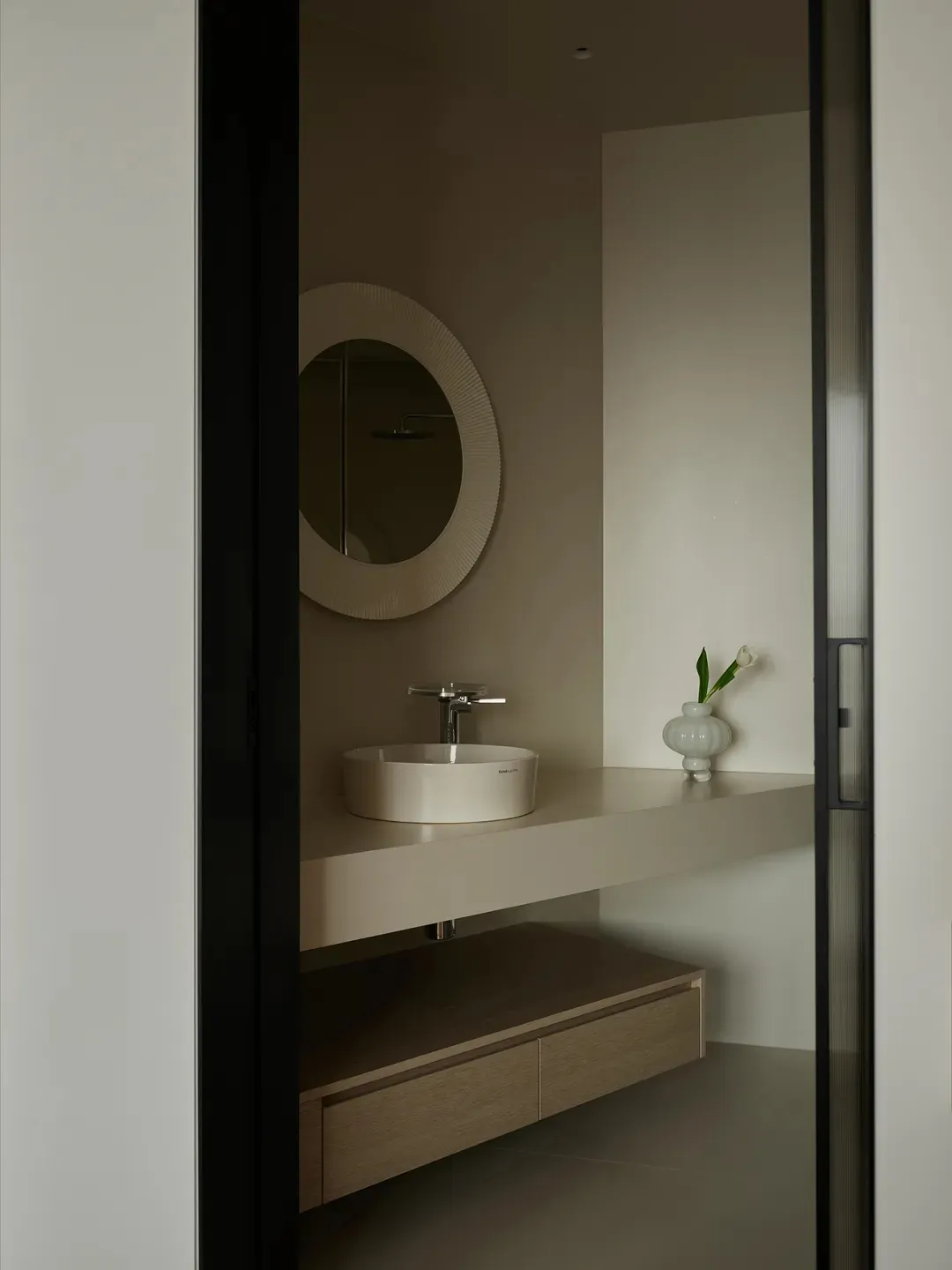 Modern minimalist bathroom with black framed glass door, white floating vanity, white sink, and white vase with green plant.