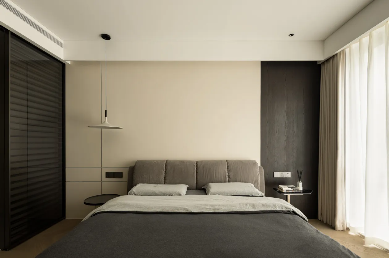 Minimalist bedroom interior featuring a bed with a upholstered headboard and grey bedding, a side table with books, and floor