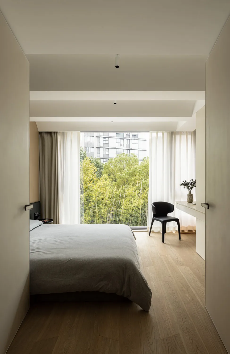 Bedroom with minimalist design, featuring a bed with wood headboard and a chair, natural light from large windows, and wooden
