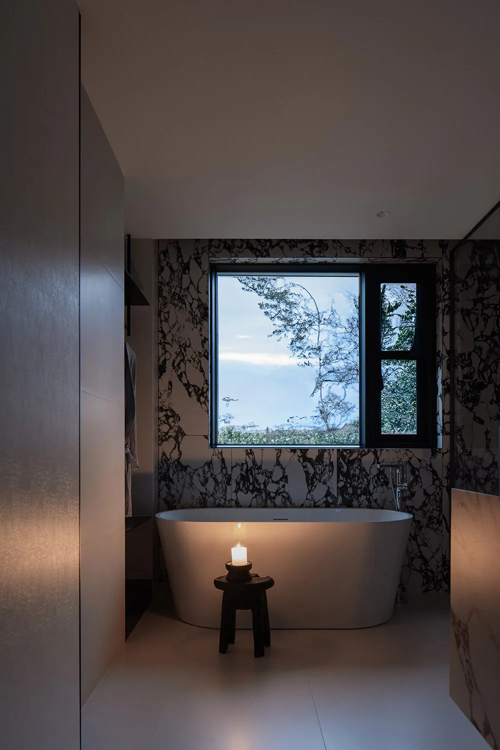 Modern minimalist bathroom featuring a freestanding tub with a candle and a view through a window with black marble wallpaper