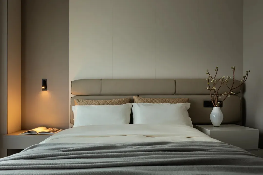A minimalist bedroom featuring a bed with a neutral-toned headboard and bedding, accompanied by a nightstand with a vase and