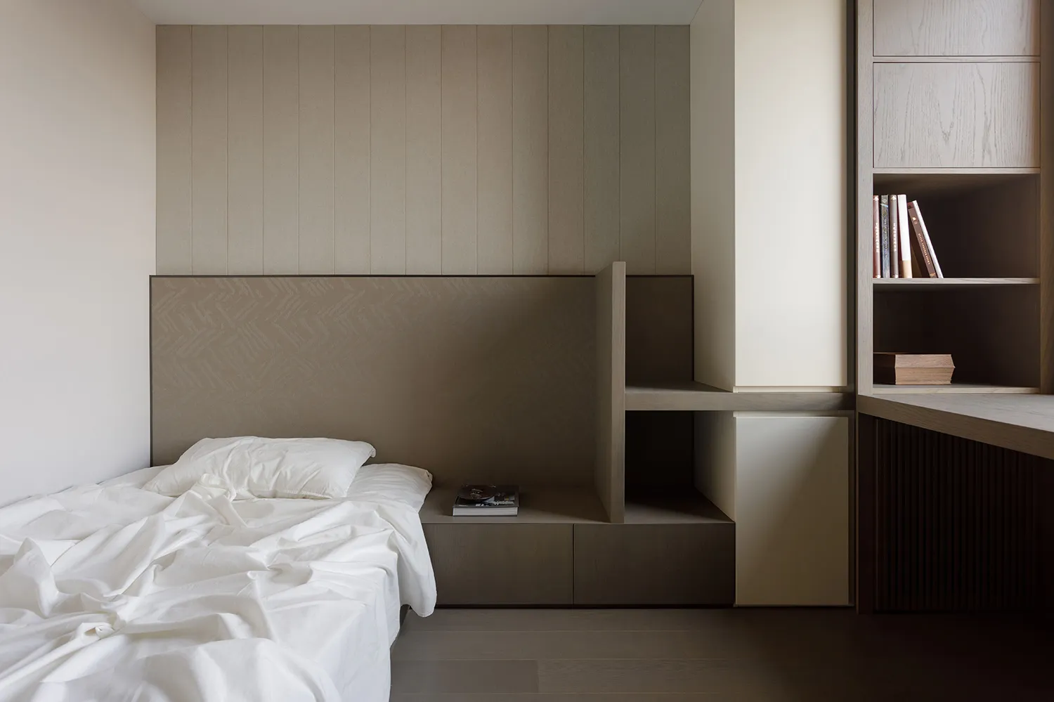 Minimalist bedroom with wooden furniture and white bedding, featuring integrated shelving and storage systems.