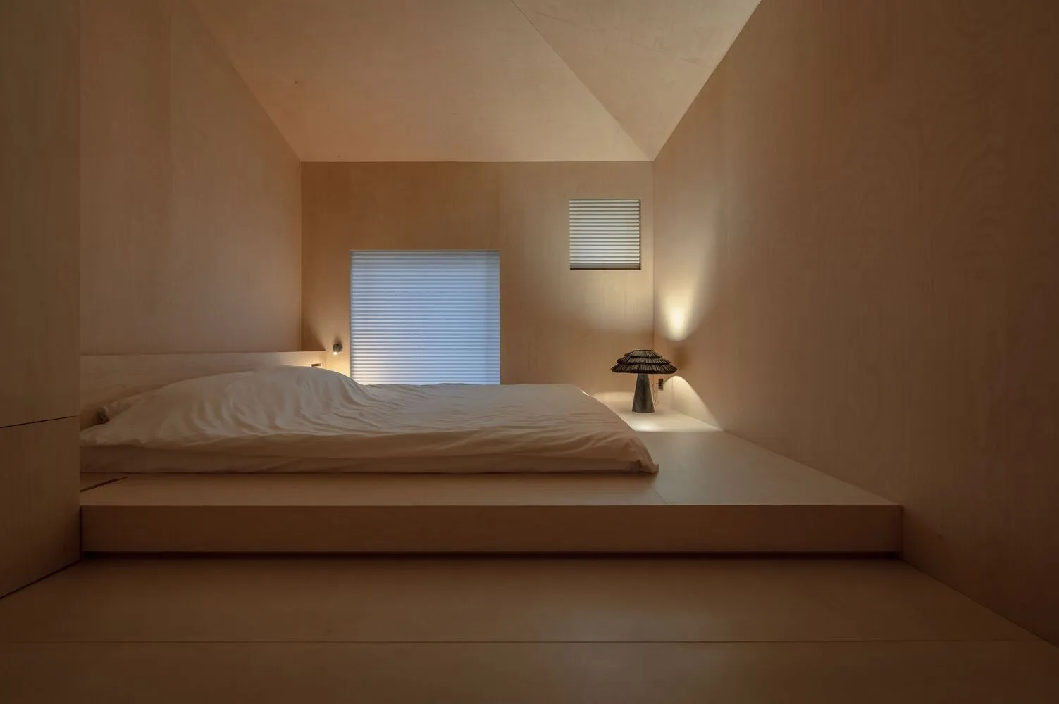 A minimalist bedroom with a wood floor, bed, and window with blinds.