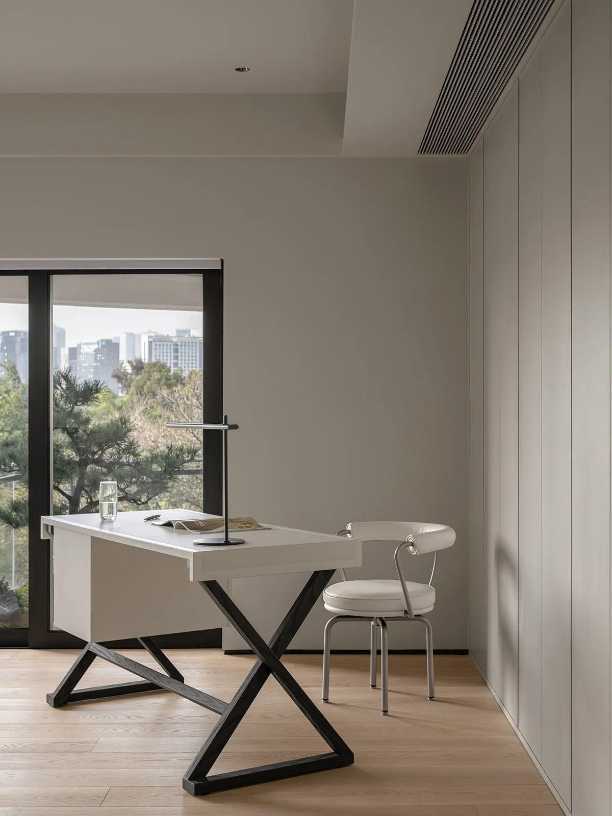 Modern home office space with sleek, minimalist desk, chair, and floor-to-ceiling windows overlooking a cityscape.