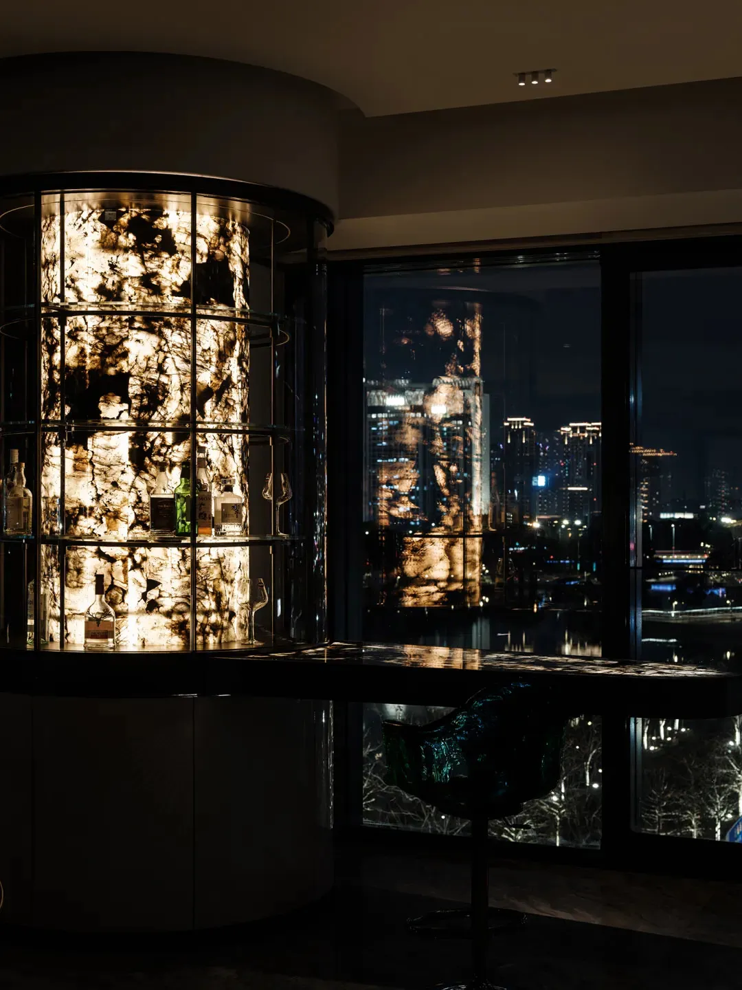 A modern bar counter featuring glass shelves and lighting, with a view of a cityscape at night.