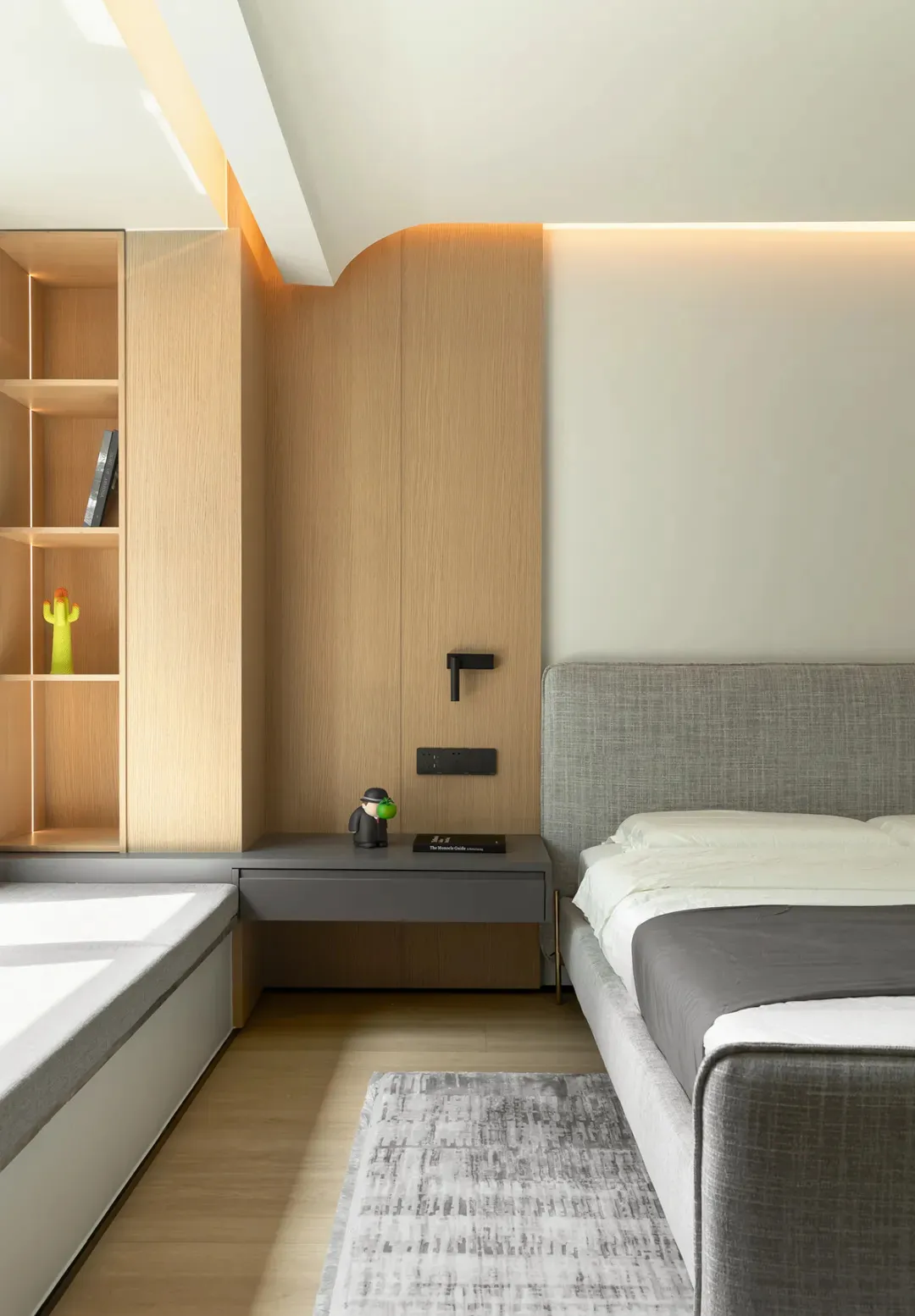 A modern bedroom featuring a grey upholstered bed with a floating nightstand and wooden shelving unit, illuminated by soft am