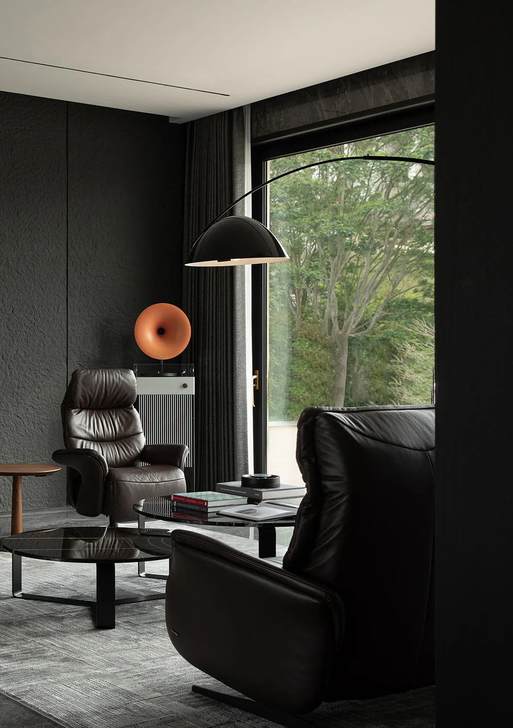 A modern living room with a black leather armchair, a coffee table with books, and a floor lamp. Glass window with trees outs