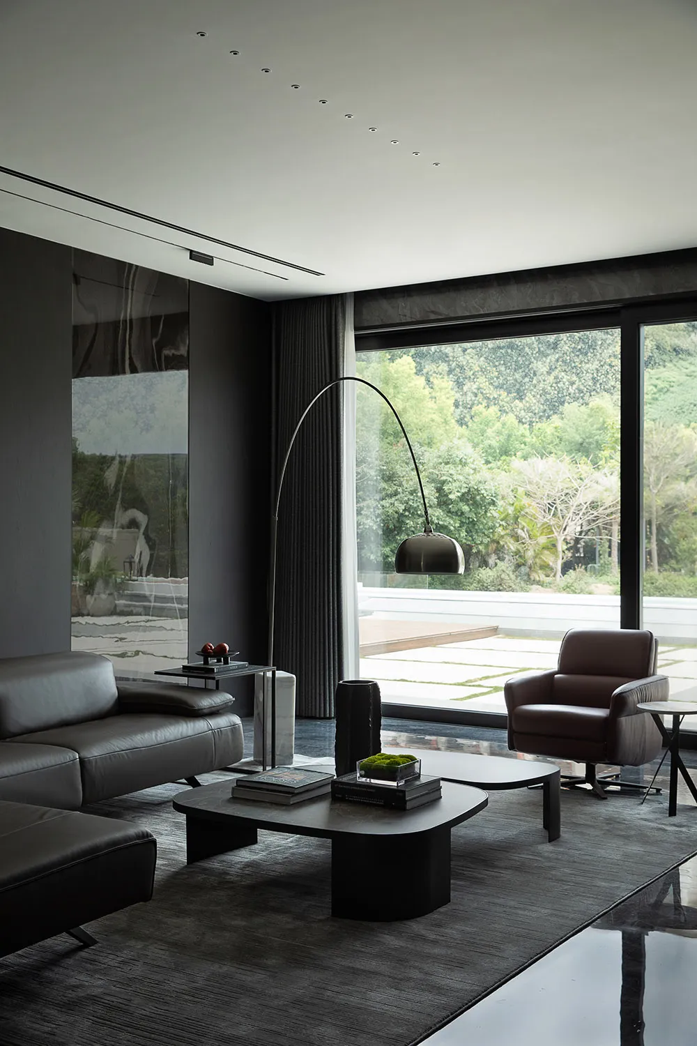 Modern living room featuring leather and glass furniture with a metal floor lamp and large windows overlooking greenery.